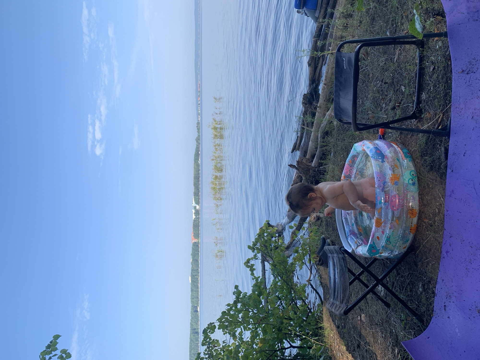 A baby is sitting in a small inflatable pool on the grassy shore of a wide river, likely the Kama River, near Chaikovsky, Russia. The baby is facing away from the camera and appears to be playing in the water. The pool is decorated with colorful cartoon sea creatures. Two folding chairs are positioned on either side of the pool, one to the left and one to the right. A purple mat is visible in the foreground, partially covering the ground. In the background, the river stretches out to the horizon, with a dense line of trees on the opposite bank. The sky is clear blue with a few wispy clouds. The scene suggests a relaxed outdoor activity on a summer day.