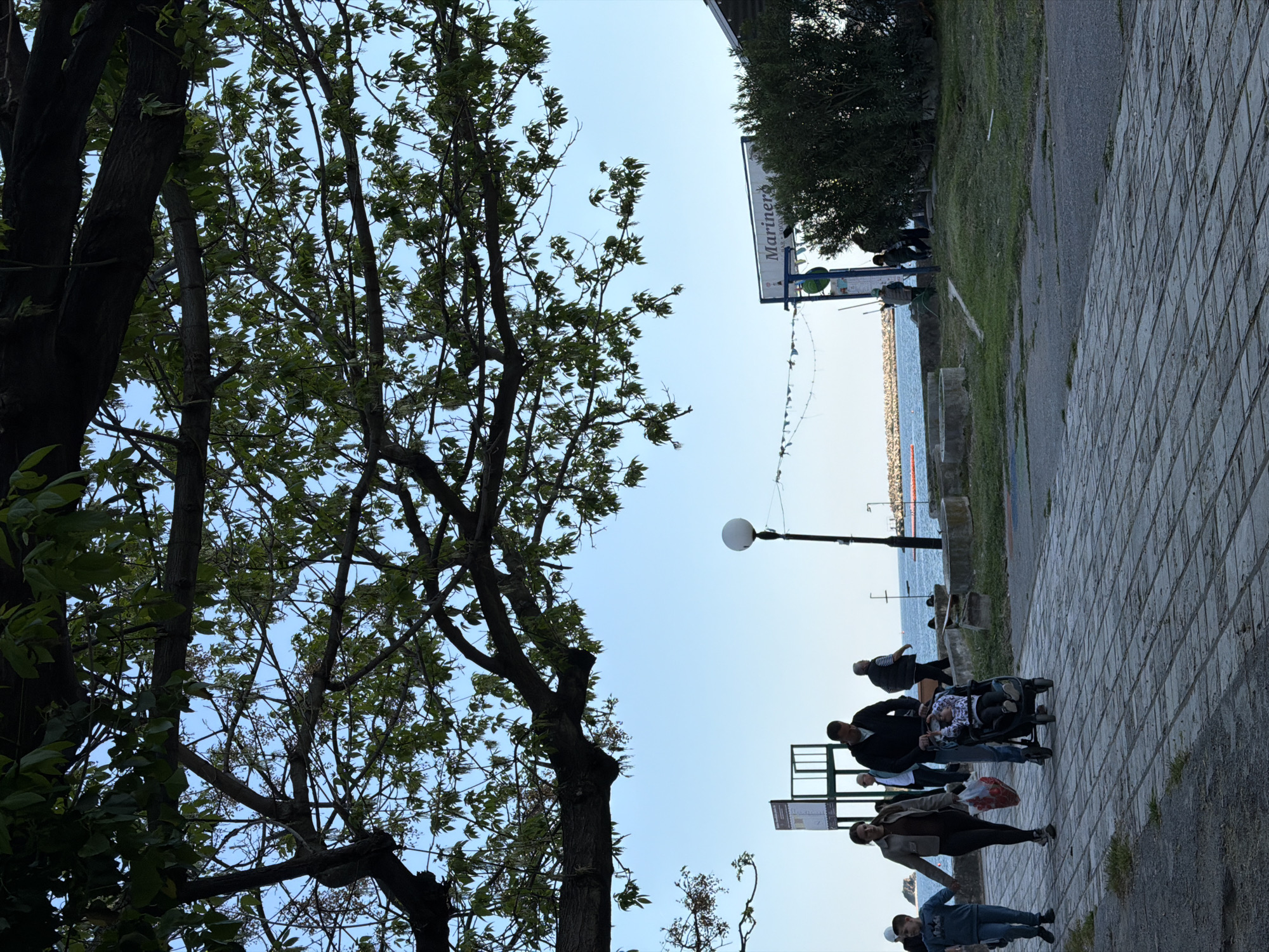 The image captures a scene along the promenade in Budva, Montenegro. A group of pedestrians walks along the paved path. To the left, trees with green foliage frame the view. In the distance, the Adriatic Sea stretches towards the horizon. A lamppost stands along the promenade. A sign reading "Marineros" is visible, partially obscured by trees.