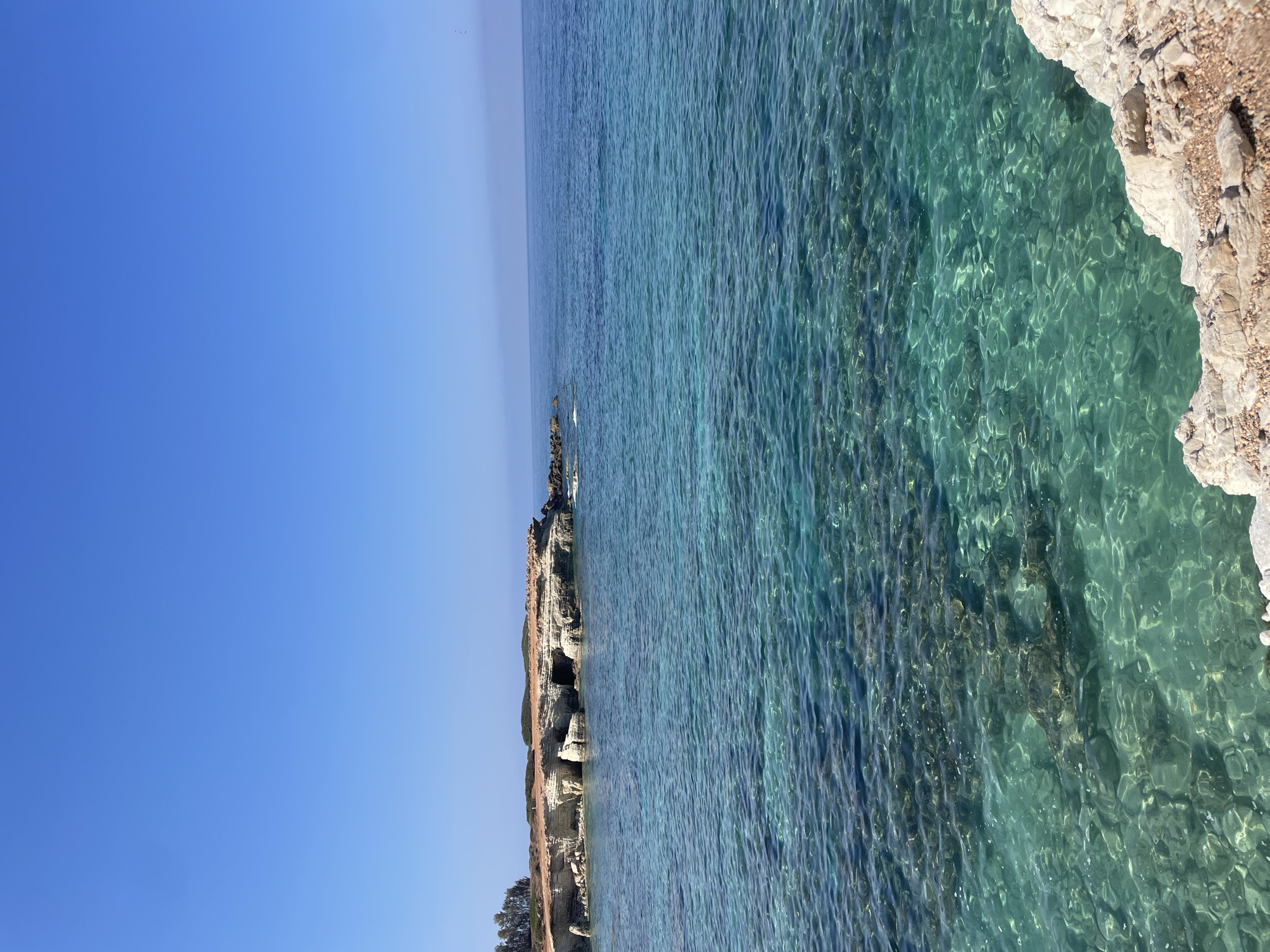 The image captures a serene coastal landscape in Peyia, Cyprus, on a clear, sunny day. The foreground is dominated by the shallow, crystal-clear turquoise waters of the Mediterranean Sea, revealing a rocky seabed beneath the surface. The water is gently rippled, with sunlight creating shimmering patterns on the ocean floor.
In the mid-ground, a rocky coastline juts out into the sea. This coastline features weathered limestone cliffs with visible caves and arches carved by the elements. A sparse scattering of dry vegetation and a few trees grow atop the cliffs. The shoreline is rugged and uneven, with exposed rocks and pebbles.
The background is characterized by a vast expanse of deep blue sky, meeting the horizon with a soft haze. The sea in the distance appears a deeper shade of blue, indicating greater depth. No people or man-made structures are visible in the frame, emphasizing the natural beauty of the location. The scene is peaceful and untouched, showcasing the geological formations and the vibrant marine environment of Peyia.