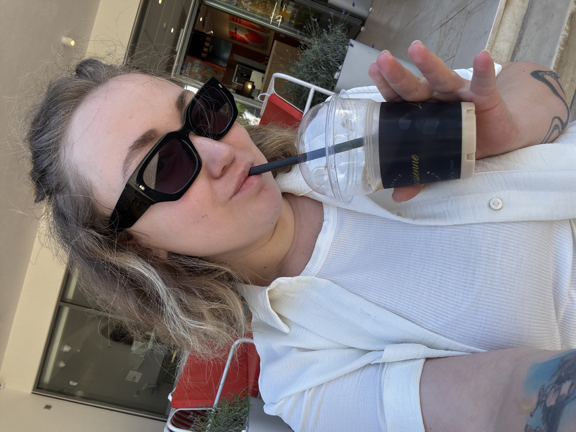 A person is taking a selfie while drinking from a clear plastic cup with a black lid and a black straw. The person is wearing black sunglasses and a white shirt. The person's hand, holding the cup, has tattoos on their forearm. The background shows a blurred outdoor scene with red chairs and plants. The person appears to be at a bar or cafe, possibly in Montenegro, given the provided location context. The time of day appears to be daytime due to the bright lighting. The overall mood seems relaxed and casual. Visible text on the cup is "L'ORIGINE PARISIENNE" and "FAITE MAISON".