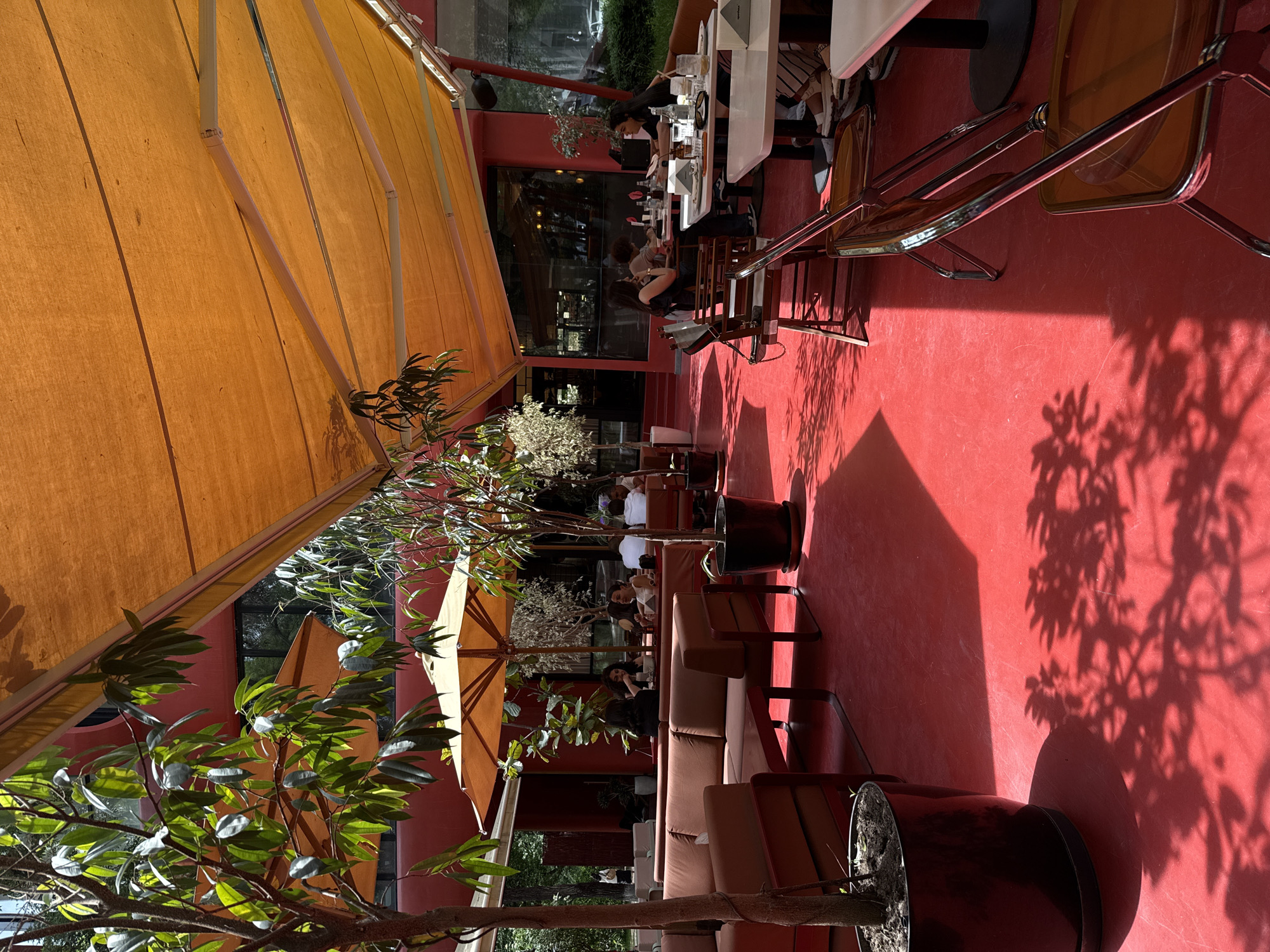 The scene is an outdoor restaurant or cafe in Yerevan, Armenia. The establishment features a large orange awning, a red floor, and a variety of seating options including chairs, tables, and couches. Several customers are present, seated at tables and engaged in dining and conversation. Decorative trees in pots are scattered throughout the outdoor space. The area is filled with strong shadows from the trees and awnings, suggesting bright sunlight.