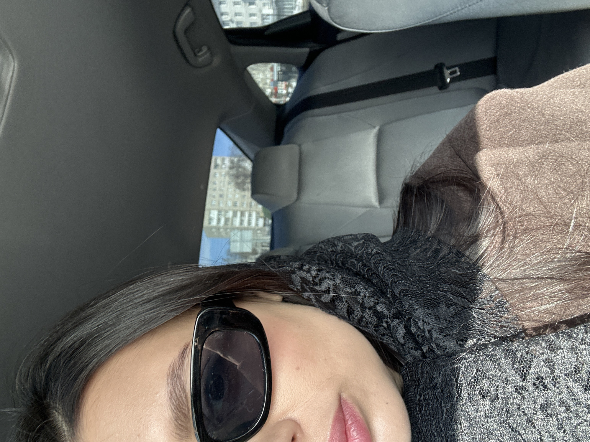 The image shows the right profile of a female individual seated in the front passenger seat of a vehicle in the city of Chaikovsky, Russia. The subject has dark hair, wears dark rectangular-framed sunglasses, and displays a fair complexion with pink-toned lips and a small mole on the right cheek below the eye. She is dressed in a dark, patterned or lace scarf and a light brown or camel-colored coat. The vehicle's interior features a light grey rear seat with a visible headrest and a black seatbelt fastened. The car's ceiling is dark grey. Through the vehicle's windows, an urban environment is visible under a clear blue sky. The background includes several multi-story residential buildings with light facades and bare deciduous trees, characteristic of a city setting.