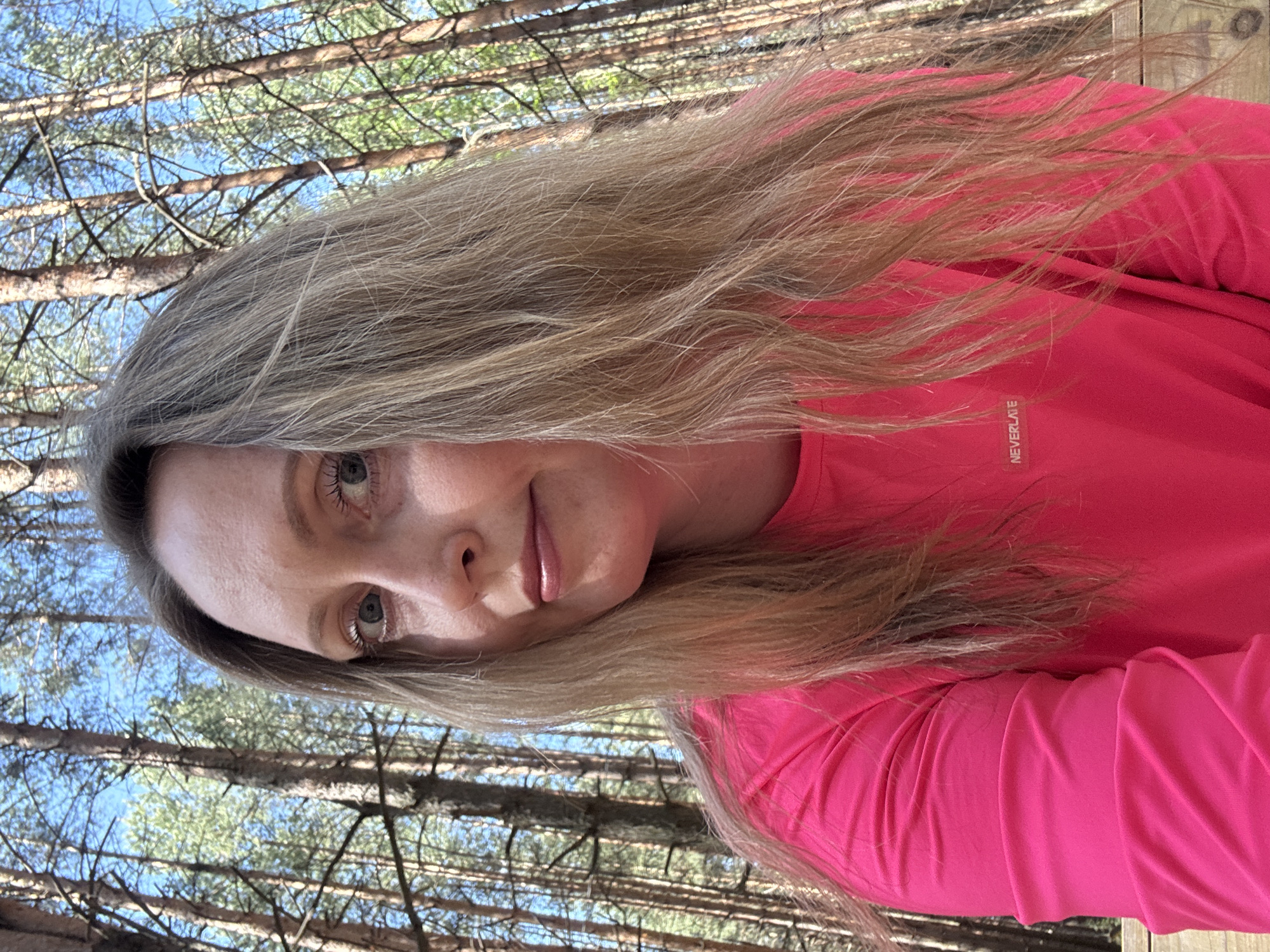 A woman with blonde hair is in a forest, possibly near Chaikovsky, Russia. She is wearing a bright pink long-sleeved shirt with the word "NEVERLATE" visible. The background shows tall trees and a blue sky.