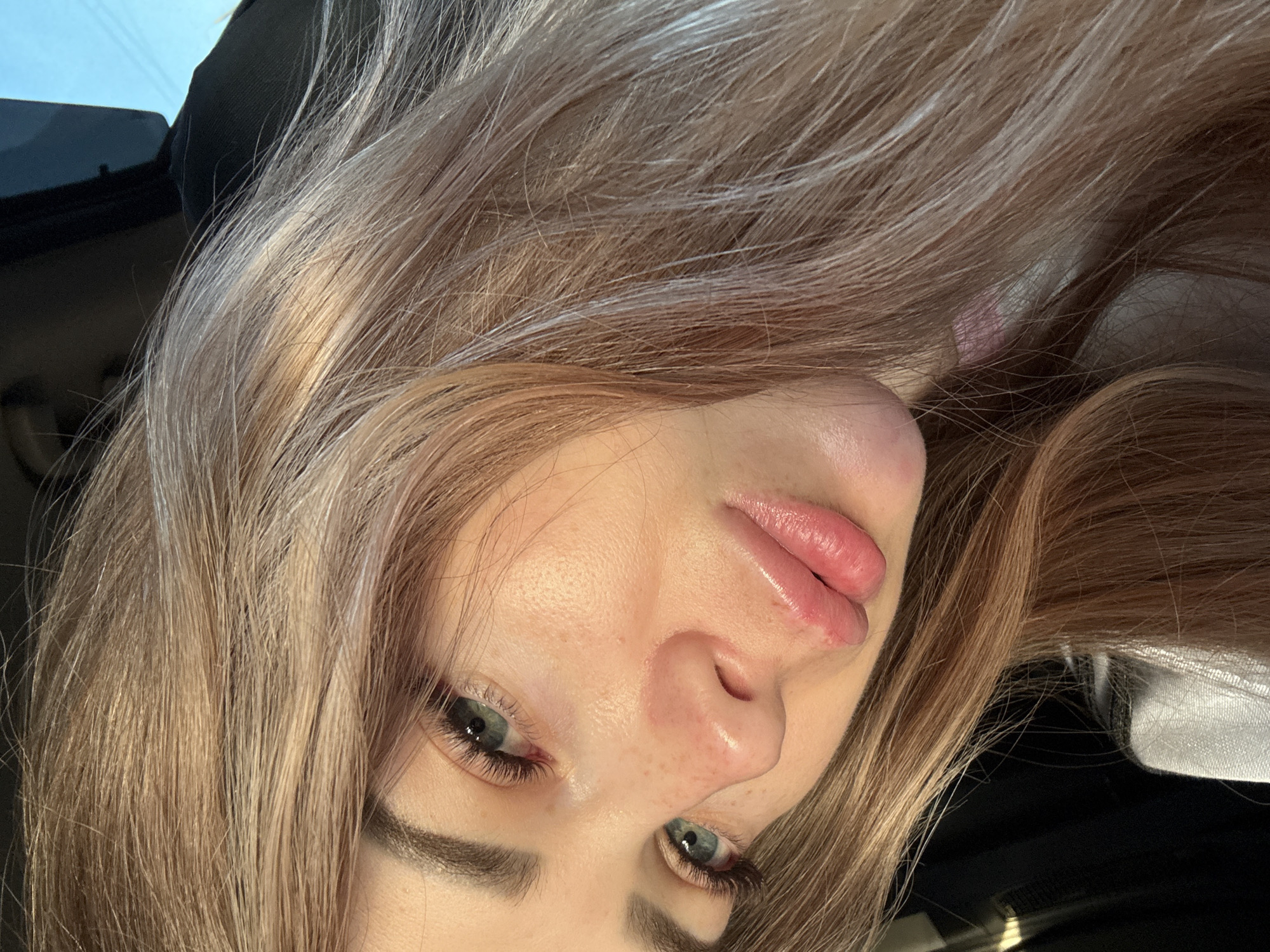 A close-up, angled portrait features a person, likely female, with long, light brown or blonde hair that cascades around her face. The image is rotated, presenting her face tilted to the side, as if looking upwards.
Her eyes, a prominent blue-green, gaze directly at the viewer. She has well-defined eyebrows and visible eyelashes, possibly enhanced with mascara or extensions. Her skin shows natural texture, with visible pores and subtle variations, and there are some faint freckles across her nose and cheeks. Her lips are full and appear glossy, with a slight pout, and a small bump or blemish is visible on her upper lip.
The hair is the most dominant element besides the face, appearing somewhat tousled and reflecting natural light with various blonde and lighter brown tones. Behind her hair, the background is largely obscured but reveals dark, soft-textured elements, likely interior upholstery, possibly of a car. A small section of a lighter, possibly sky-blue, area is visible in the top left corner, suggesting a window.
The lighting is bright and natural, highlighting features on her face and hair. The overall impression is an intimate, informal portrait, possibly a selfie, captured in daylight within an indoor setting, potentially a vehicle. No visible text is present.