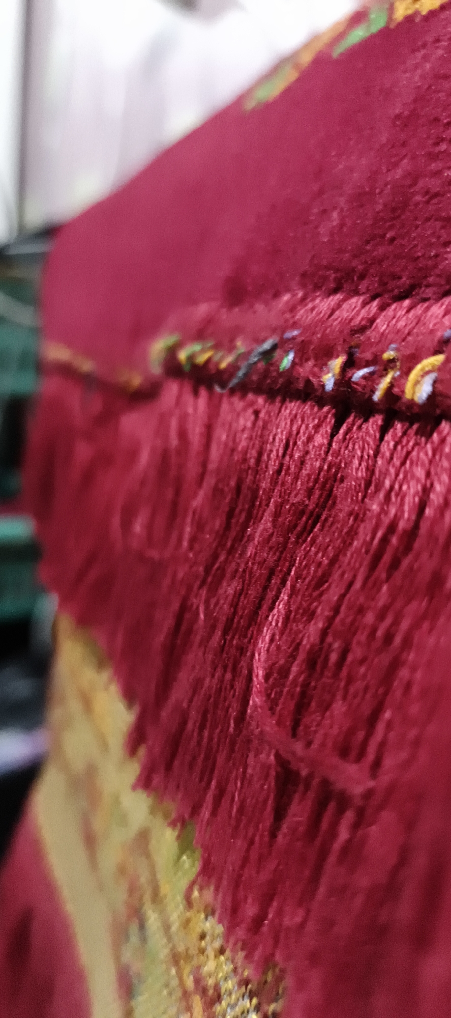 The image shows a close-up view of a red fringed textile, likely a carpet or tapestry. The fringe is made of many thin threads, and at the top edge of the fringe, there is a decorative band with multicolored threads in yellow, green, blue, and white. The textile appears to be on a surface, and a portion of another textile with a gold and yellow pattern is visible underneath and to the left. The background is blurred and out of focus, with hints of white and green. There are no people or specific landmarks visible in the image. Without additional context, it is impossible to determine the specific location in Muaro Jambi, Indonesia.