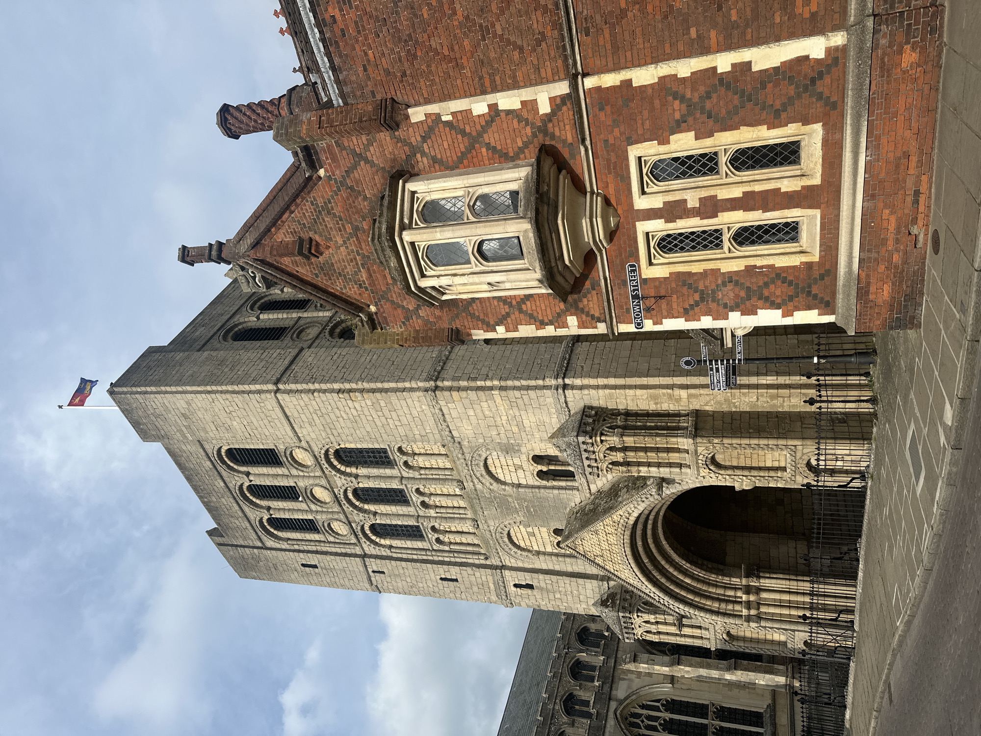The image shows St Edmundsbury Cathedral in Bury St Edmunds, West Suffolk, United Kingdom. The cathedral tower, constructed of light-colored stone, rises prominently against a partly cloudy sky and displays a flag on its peak. The tower has multiple levels of arched windows. Adjacent to the tower is a red brick building with decorative patterns. A bay window and adjacent windows are visible on the brick building, and a street sign labeled "Crown Street" is mounted on the wall. Wrought iron fencing is present in front of the cathedral entrance. The foreground includes part of a paved area.
