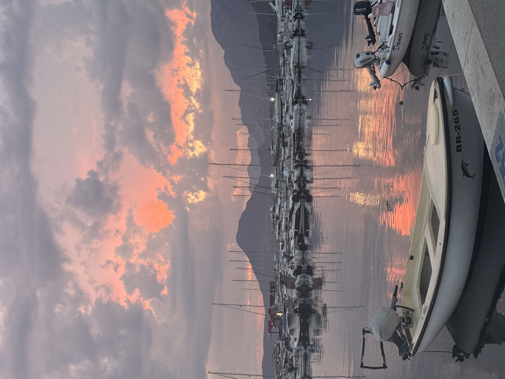 This is a medium shot, captured from a slightly elevated position, of a marina in Bar, Montenegro during sunset. The sky is filled with dramatic clouds, tinged with vibrant orange and pink hues, reflecting on the calm water. Numerous sailboats and smaller motorboats are docked in the marina, their masts and hulls creating a dense, textured silhouette against the colorful sky and the dark, mountainous landscape in the background.
In the foreground, two small white motorboats are prominently featured. The boat on the left has a dark hull trim and the inscription "BR-265" along with a dolphin silhouette. It is equipped with a light-colored engine cover. The boat on the right, partially visible, has the name "Damir" written on its side. Both boats are bobbing gently on the water. The surface of the water acts as a mirror, reflecting the boats, their reflections, and the fiery sky above. The overall impression is one of tranquility and the beauty of nature during a coastal sunset.
