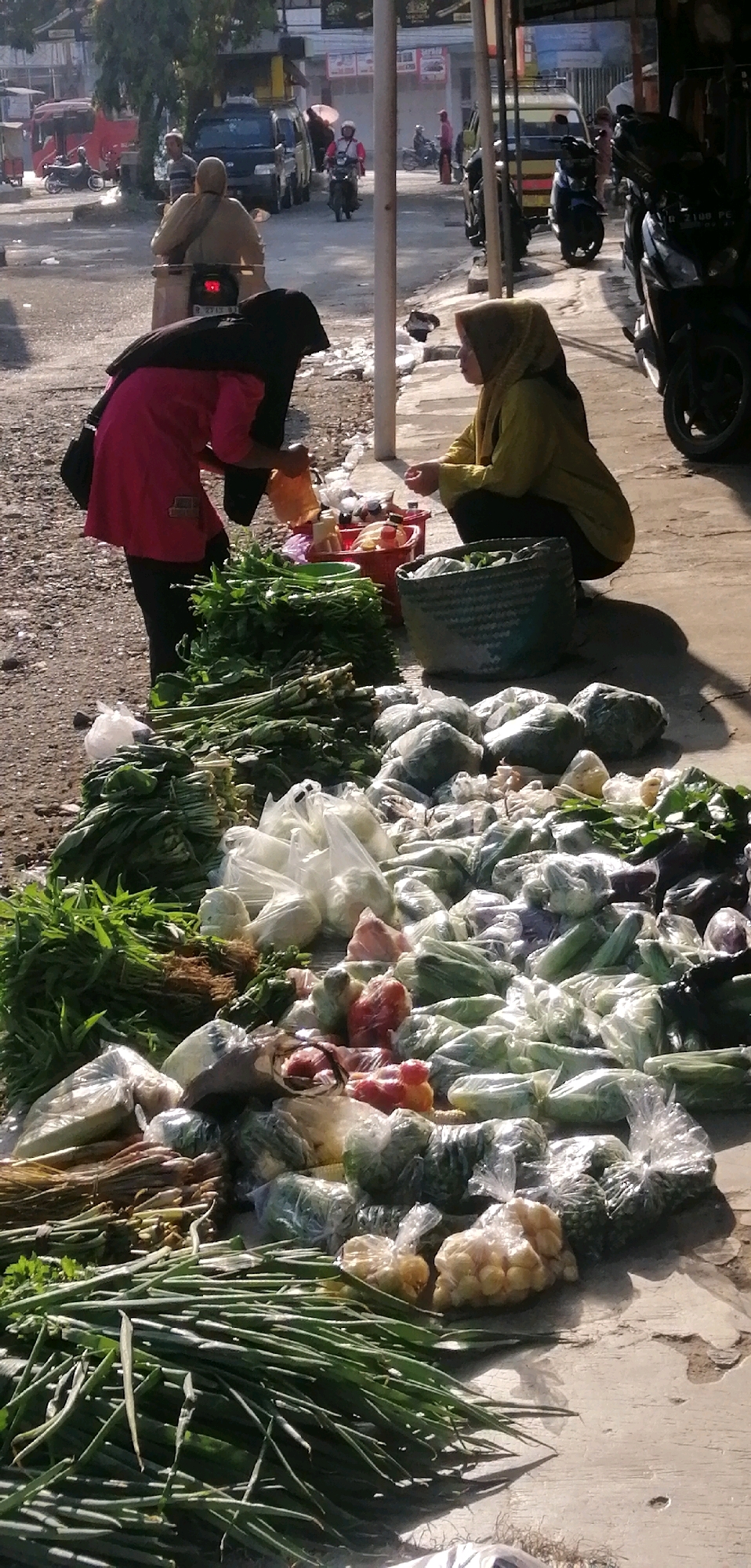 In Ajibarang, Indonesia, a street market scene unfolds with two women engaged in commerce. One woman, dressed in a pink top and black headscarf, is bent over, interacting with produce. The other woman, in a yellow headscarf and top, is crouching, seemingly having a transaction. Piles of fresh vegetables, including bundles of green onions, leafy greens, and various other produce wrapped in plastic bags, are displayed on the ground and in a woven basket. The background shows a street with vehicles, including motorcycles and a van, suggesting a typical urban environment. Sunlight casts shadows, indicating it is likely daytime.