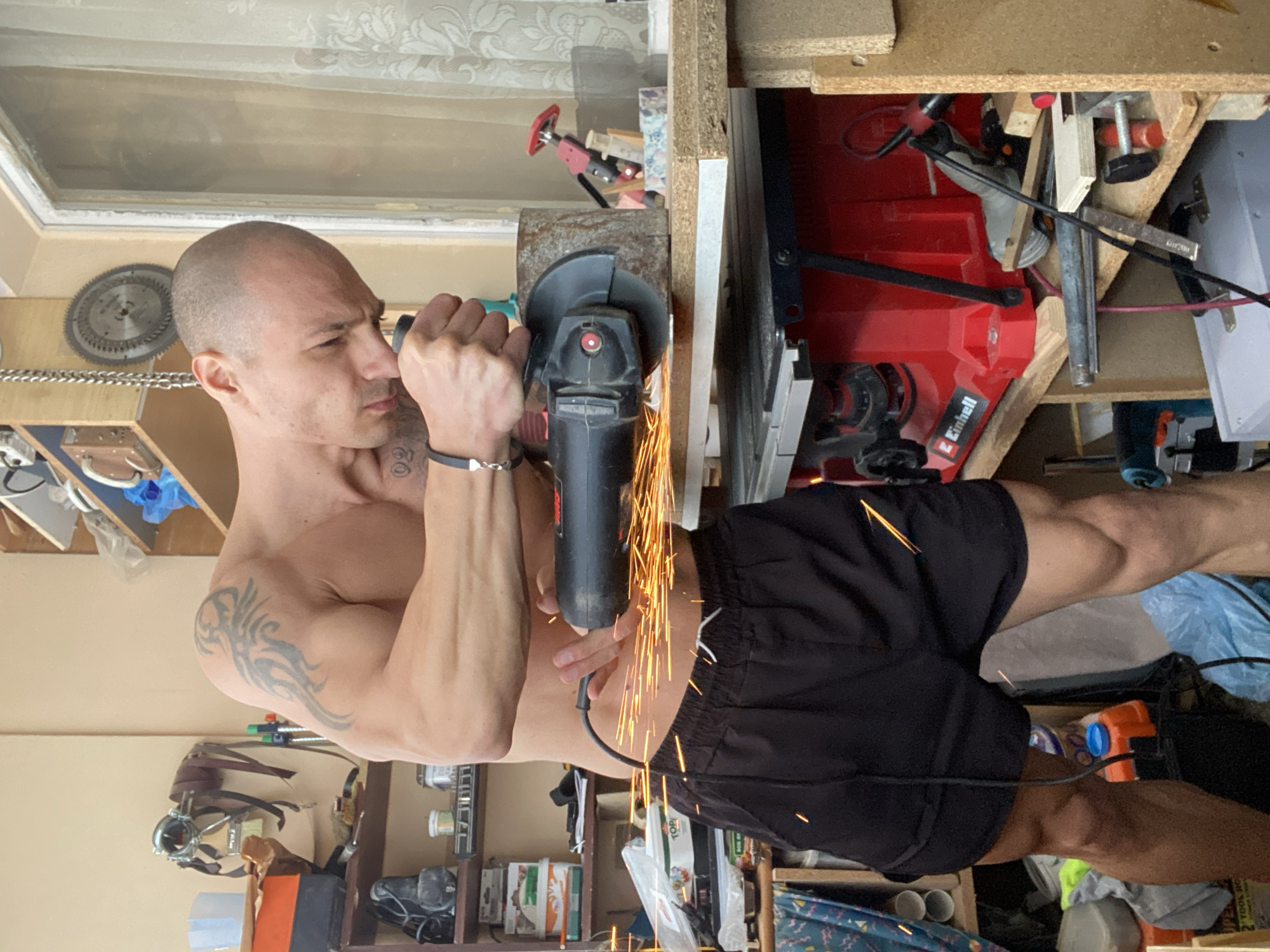 A shirtless man with a shaved head and visible tattoos is using a grinder to cut a metal object. Sparks are flying from the cutting point. He is wearing black shorts. The background contains a cluttered workshop environment with shelves holding various tools and materials, including a table saw. There's a window in the upper left. The image appears to be rotated. There is no information available to determine if this photo was taken in the city of Pleven, Bulgaria.
