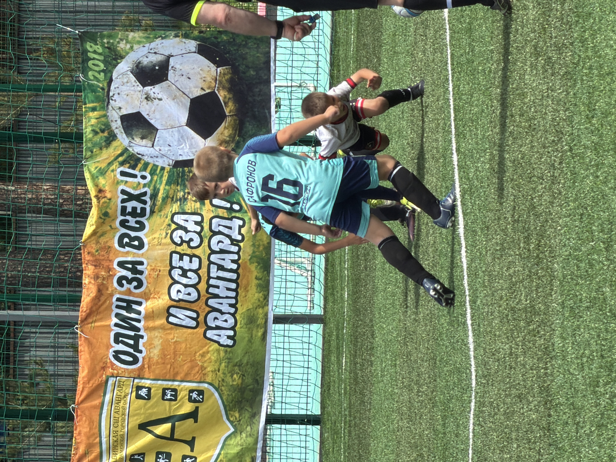 This is a vertically oriented, close-up photograph of two young boys playing soccer on a bright, sunny day in Bykovo, Russia. The boys are dressed in soccer uniforms. One boy, wearing a light blue and navy jersey with the number 16 on the back and the name "САФРОНОВ" above it, is in the foreground. He is looking towards the right of the frame with a determined expression. The other boy, in a white and red jersey and black shorts, is in the background, seemingly in motion.
Behind the boys is a large banner with a soccer ball graphic and text in Russian. The banner reads "2018" and then "ОДИН ЗА ВСЕХ !" which translates to "One for all!". Below that, it says "И ВСЕ ЗА АВАНГАРД!" or "And all for Avangard!". There are also some smaller graphics and text at the bottom of the banner, including "ЛЬИНСКАЯ СШ АВАНГАРД" and "РАМЕНСКИЙ ГОРОДСКОЙ ОКРУГ", likely referring to the name of the sports school and the district.
The boys are on a green artificial turf soccer field, demarcated by a white line. A green mesh fence runs along the left side of the frame. The lighting suggests it is midday, with strong sunlight casting shadows. The overall scene depicts a youth soccer match or practice, filled with youthful energy and sportsmanship.