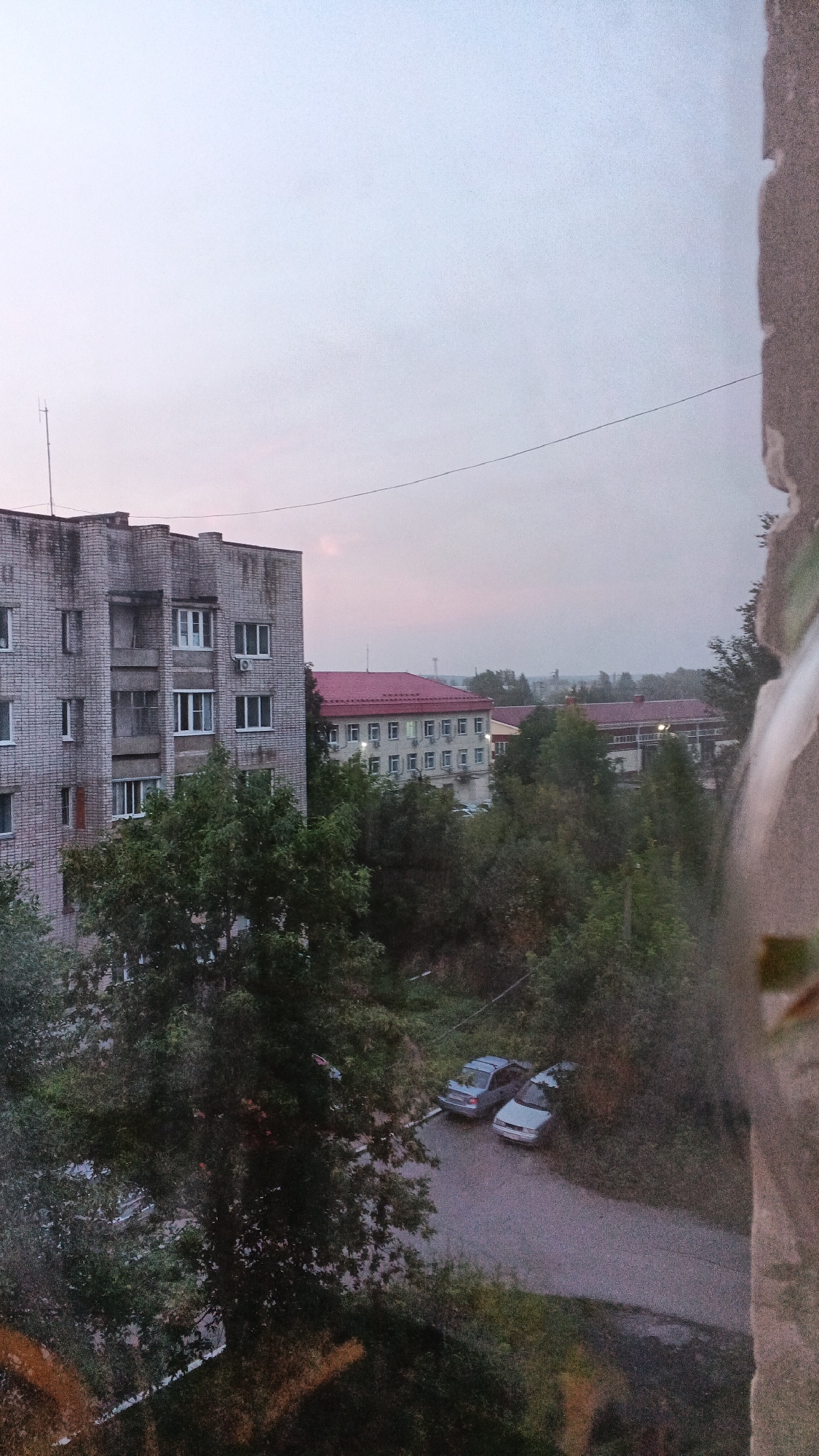 The image depicts a view from a high vantage point, likely a window, looking out onto an urban landscape under an overcast sky. In the foreground, a gray, multi-story brick apartment building is visible on the left. The building shows signs of age with some staining on its facade. To the right of this building, a cluster of trees and foliage partially obscures the view of other structures.
Further into the scene, a longer, lighter-colored building with a red tiled roof is prominent. This building appears to be institutional, possibly a school or administrative facility, with a uniform row of windows and air conditioning units visible. In front of this building and beyond the trees, a road or paved area is visible, wet as if from recent rain. Two cars, a silver sedan and a lighter-colored car, are parked on this wet surface. The background is filled with more trees and suggests a residential or semi-urban environment. The overall lighting suggests it is either early morning or late evening, with the sky exhibiting subtle hues of pink and gray. The location is identified as the city of Izhevsk, Russia.