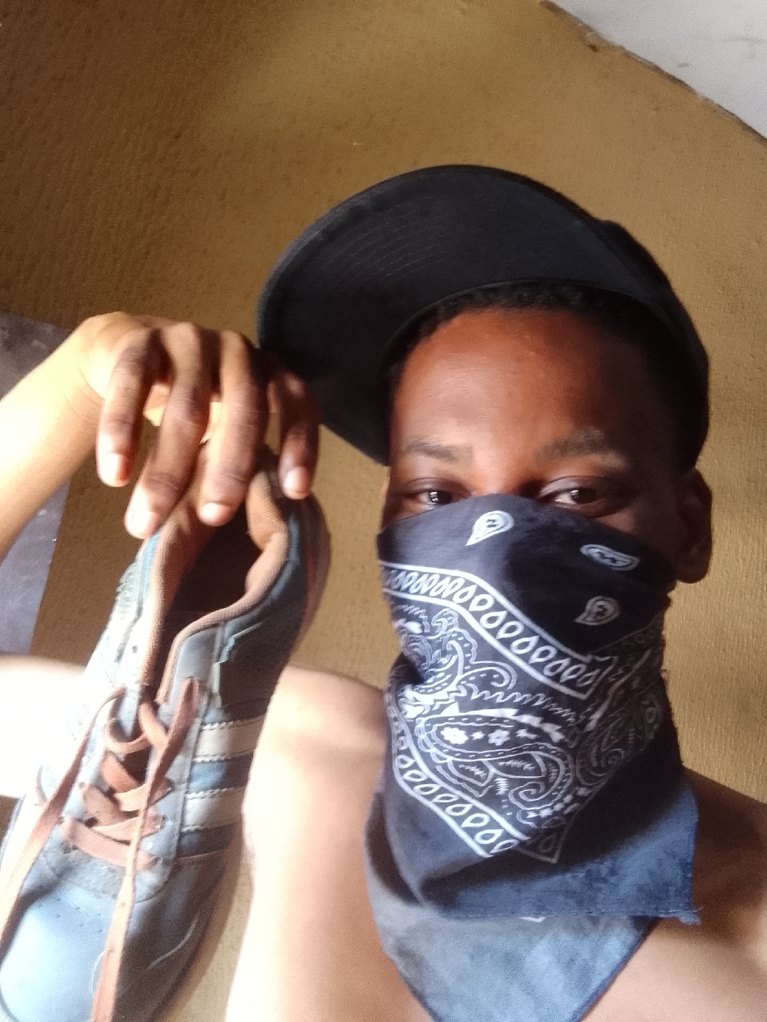 A person is depicted in a medium close-up shot, looking directly at the camera. They have dark skin and are wearing a black baseball cap. A black bandana with a white paisley pattern covers their nose and mouth, obscuring most of their face except for their eyes and forehead. With their left hand, the person is holding up a single sneaker, presenting it towards the viewer. The sneaker is grey or blue with brown laces and features three distinct stripes on its side, characteristic of the Adidas brand. The background is a textured, warm-toned wall, likely painted yellow or light brown, suggesting an indoor setting. The lighting is bright and even, indicating daytime. No explicit text is visible, but the shoe's design strongly implies its brand.