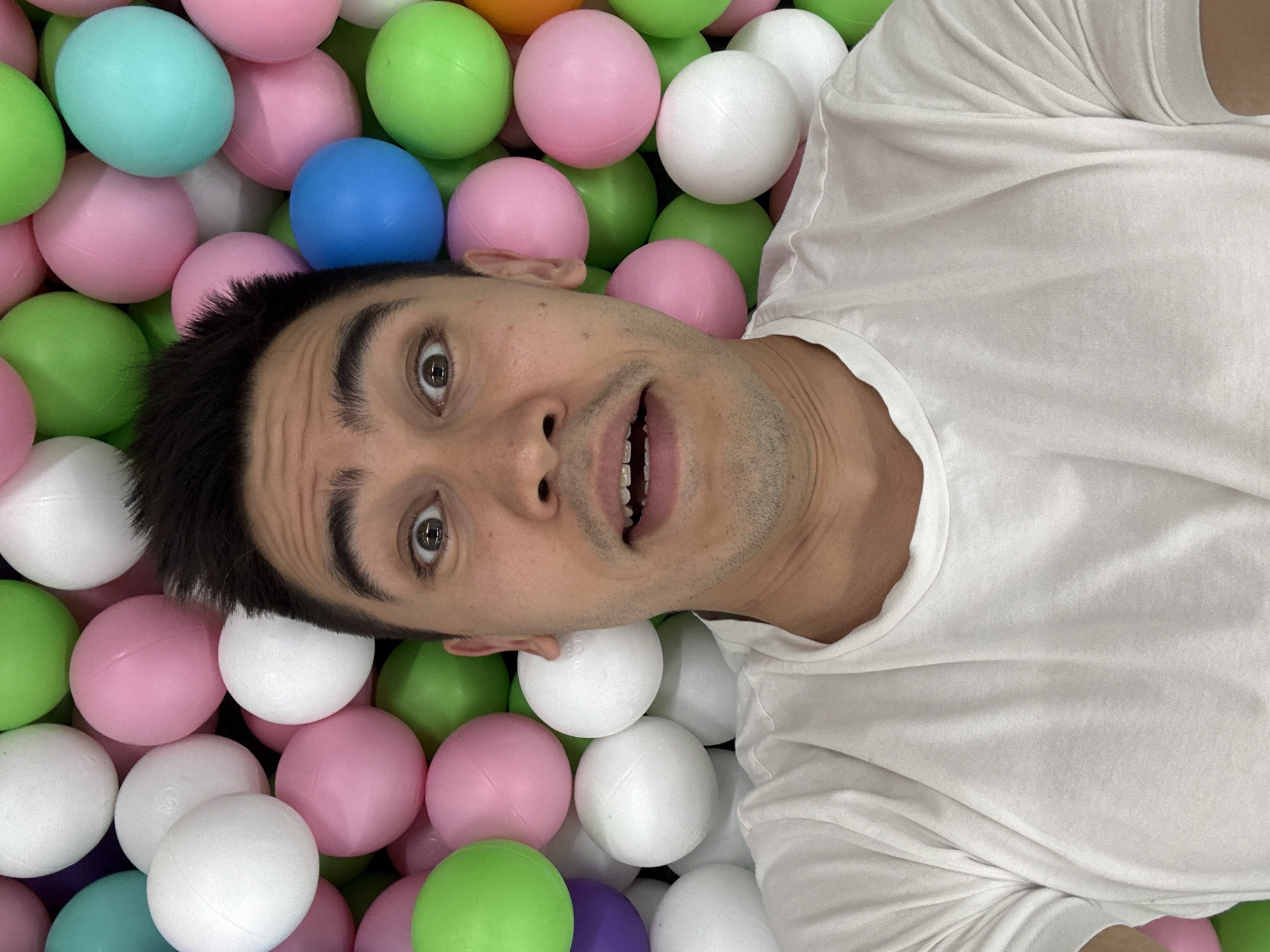 An overhead shot captures a male individual lying submerged in a colorful ball pit in Kazan, Russia. His head and upper torso are visible, with his body oriented diagonally across the frame. He has short dark hair, light brown eyes, and visible stubble on his chin and cheeks. His eyes are wide open, and his mouth is agape, revealing his upper teeth, conveying an expression of surprise, amusement, or playfulness. He is wearing a plain white t-shirt, which is partially covered by the balls.
The ball pit is filled with numerous plastic spheres in various hues, predominantly pastel pink, light green, white, and light blue. Hints of purple and orange balls are also visible. The balls are densely packed around the person, creating a vibrant, playful environment. The lighting appears even, suggesting an indoor setting like a recreational center or amusement park. There is no visible text in the image.
