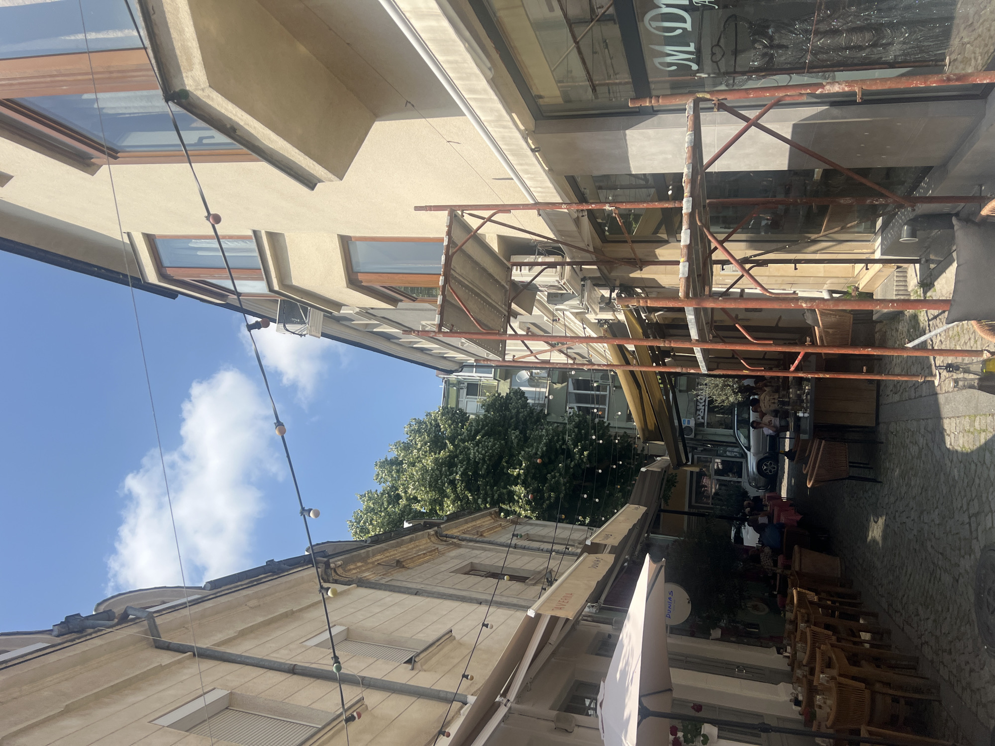 The image captures a vibrant, sunny pedestrian street scene, likely in a European city, with buildings lining both sides. The sky is bright blue with scattered white clouds, indicating daytime.
**Subjects and Scene:** On the left, a multi-story building with light-colored walls and brown-framed windows, including what appear to be skylights, stands prominently. String lights with small bulbs are strung across the street, connecting to this building and the one opposite. On the right side of the street, another building is partially obscured by extensive, rusty metal scaffolding, suggesting ongoing construction or renovation. This building features a large glass storefront, reflecting parts of the street. Further down the street, lush green trees are visible, adding a natural element to the urban environment.
**Activity:** People are seated at numerous outdoor tables and chairs, predominantly on the right side of the street. They appear to be patrons of cafes or restaurants, enjoying drinks or meals in the open air. A silver car is parked further down the street, suggesting some vehicular access, although the immediate area seems dedicated to pedestrian activity and outdoor dining.
**Notable Details:** The atmosphere is bright and lively, characteristic of a bustling urban center. The outdoor seating, decorative lights, and mixture of architectural styles contribute to a charming European street ambiance. The scaffolding introduces an element of transition or maintenance within the established setting. Air conditioning units and a satellite dish are visible on the buildings, indicating modern amenities.
**Visible Text:** Several pieces of text are visible. On the storefront behind the scaffolding, "MD" is part of a larger sign, possibly "MDIVA". On various awnings and an umbrella, "TAVERNA", "JONAS", and "DUNIA'S" can be read. Near the parked car, a sign with partial text "PAKC H" suggests "PARKING".