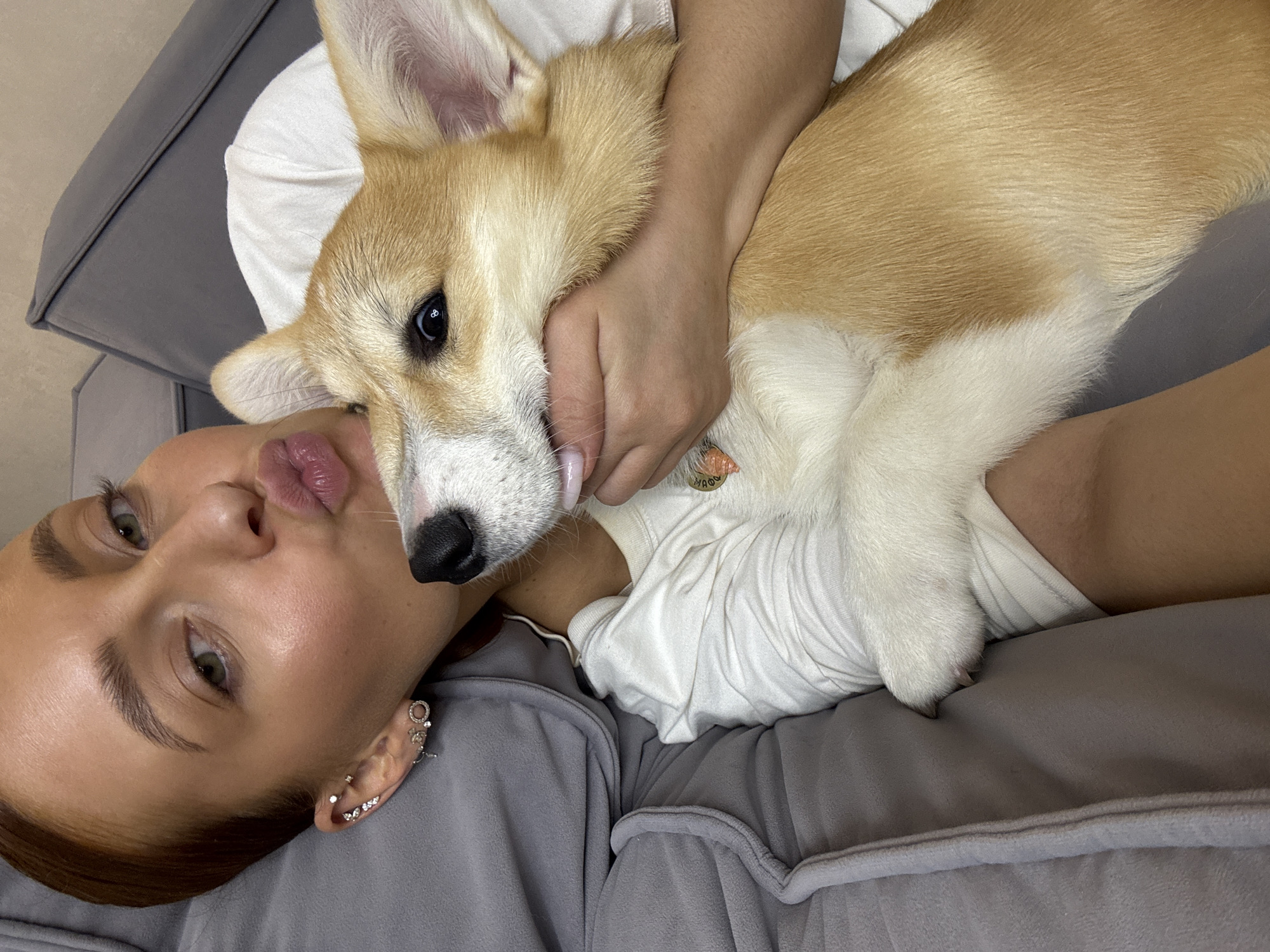 A young woman with light skin, dark eyebrows, and green eyes lies on a grey, soft-textured couch or bed, taking a selfie. Her lips are playfully pursed in a 'kissy face' expression. She is wearing a white top and her right ear is adorned with several small, shiny earrings. Cuddled closely beside her, with its head resting on her shoulder, is a fawn and white Pembroke Welsh Corgi. The woman's arm is wrapped around the dog, indicating affection and comfort. The dog has dark, alert eyes and appears calm and content in her embrace, its fur looking soft and well-groomed. A small, round tag is visible on the dog's collar, bearing faint text that appears to read "ΦΛΩΡΑ" (Flora). The scene captures a cozy, affectionate moment between a pet owner and her dog, likely indoors during the daytime in a domestic setting, with a warm and relaxed atmosphere. The location context is Chaikovsky, Russia.