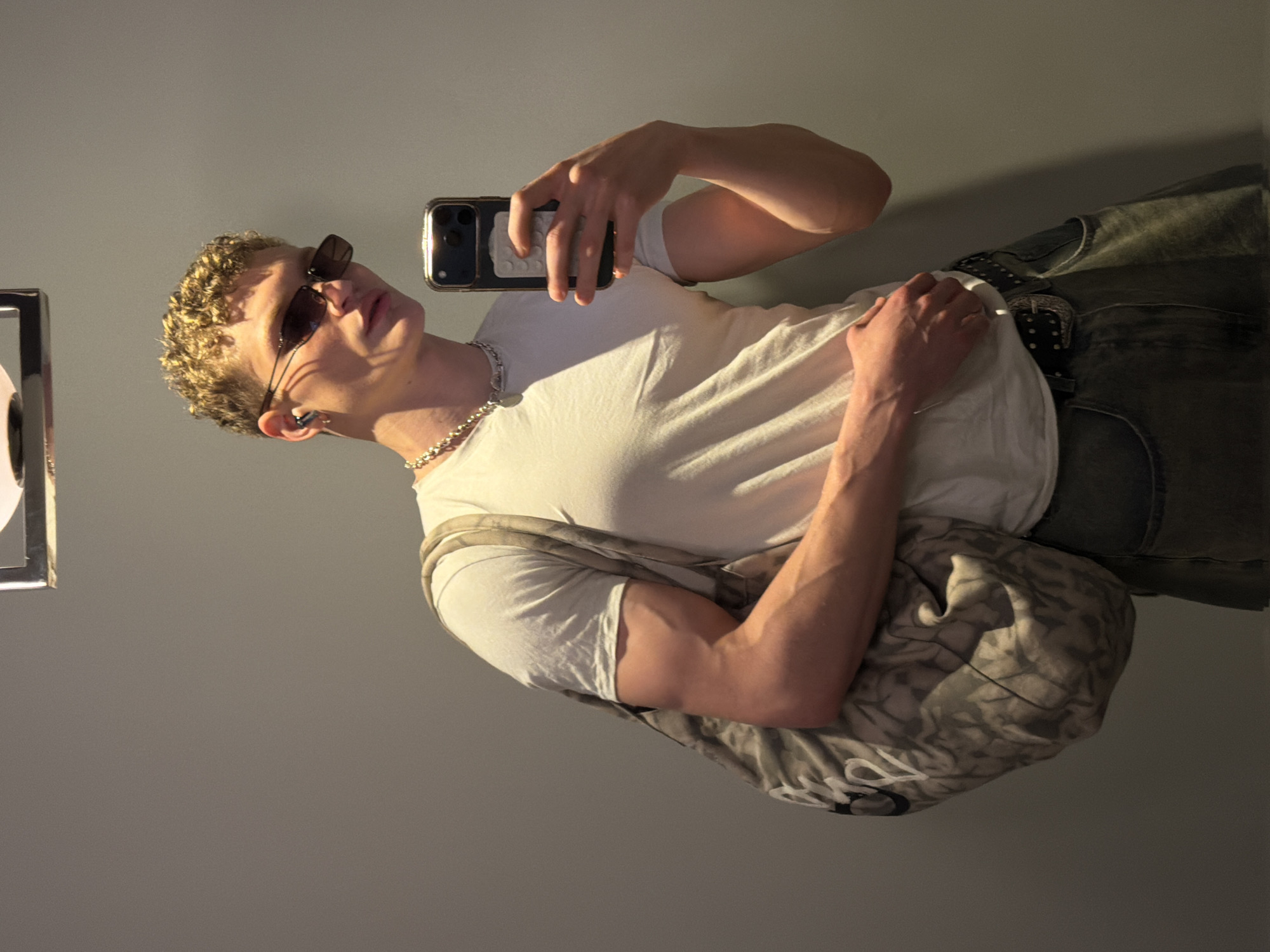 A young, fair-skinned male takes a selfie. He has short, curly blonde hair and wears sunglasses, a silver chain, a white t-shirt, dark jeans with a studded belt, and carries a gray patterned shoulder bag. He holds a smartphone in his right hand, capturing the image. The background consists of a plain gray wall, with a framed object on the left side. There is no indication of the location being in London, United Kingdom.