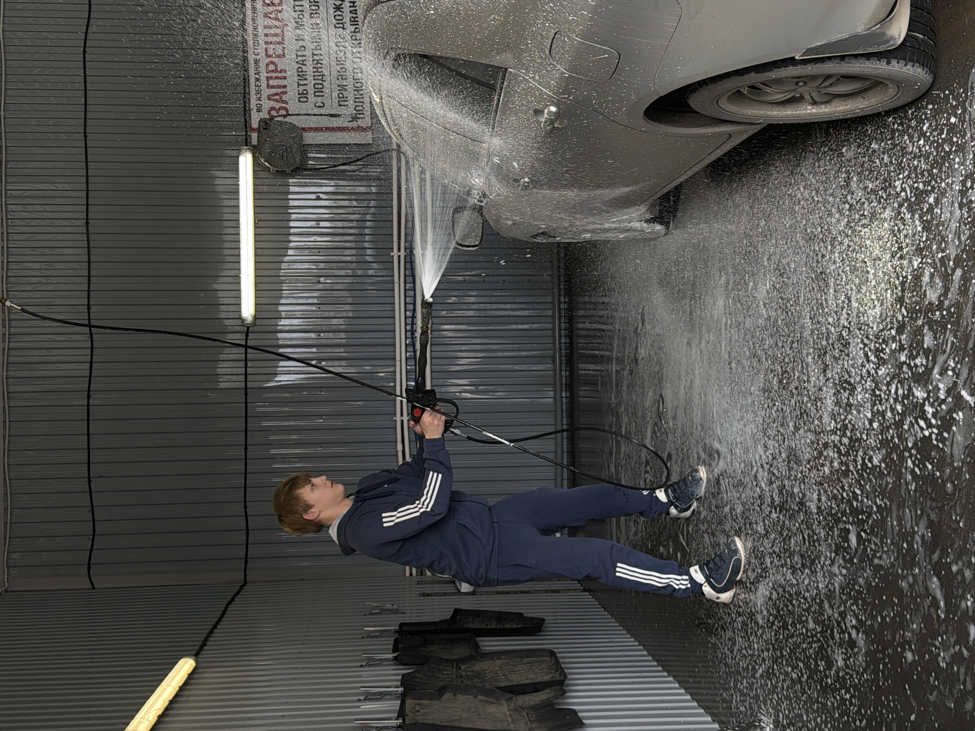 Here's a detailed description of the image:
**Scene Overview:** The image shows a person washing a car in what appears to be a car wash bay. The overall environment is industrial and functional.
**People:** A young adult male is depicted in the act of washing a car. He is wearing a navy blue tracksuit with white stripes on the sleeves and pants. He is wearing dark sneakers with white trim.
**Subjects:** The primary subject is the partially visible silver-colored car being washed.
**Objects:**
* A high-pressure water hose and nozzle is in use.
* Car mats are stacked on the floor near the person.
* Fluorescent lights illuminate the bay.
* A sign with Russian text is mounted on the wall.
**Actions:** The person is holding the water hose and directing a stream of water towards the car, seemingly in the process of cleaning it.
**Interactions:** There is an interaction between the person, the water hose, and the car, specifically aimed at cleaning the car's exterior.
**Environment:** The car wash bay has corrugated metal walls and appears to be partially enclosed. The floor is wet and covered in soap suds. The scene is brightly lit.
**Text Analysis:** A sign is visible in the background with text that includes: "ВО ИЗБЕЖАНИЕ СТОЛКНОВЕНИЯ ЗАПРЕЩАЕ" ("To avoid collisions prohibited").
**Location:** Given the Russian text on the sign, it is potentially Chaikovsky, Russia.