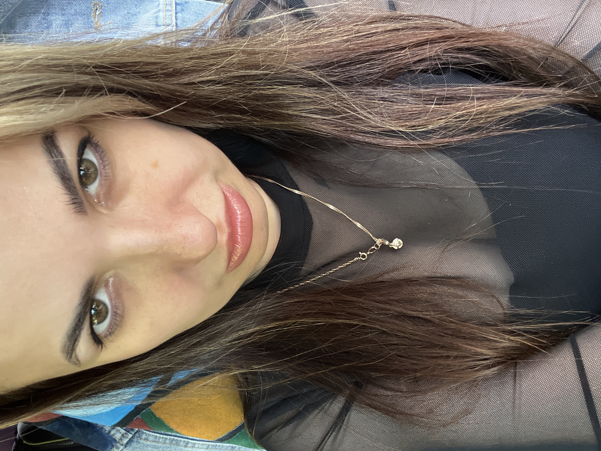 A woman is facing the camera. She has long brown and blonde hair, and her skin is fair. She is wearing a black mesh top and a gold necklace. She appears to be indoors.
