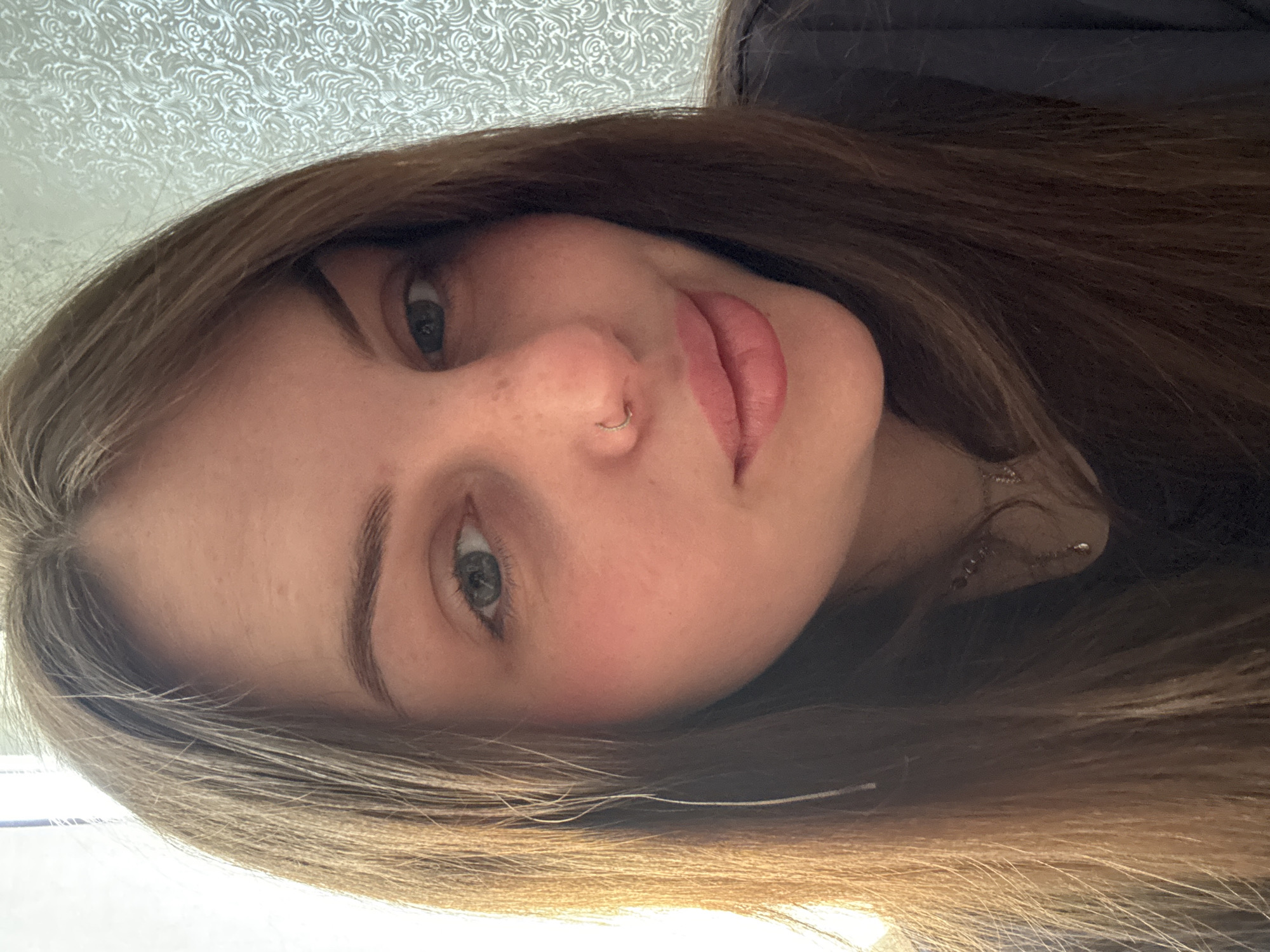 A close-up, slightly angled portrait captures a young woman with light brown hair. Her eyes are a blend of blue and green. She has a fair complexion with slight freckling around her nose. A silver nose ring is visible on her left nostril. Her lips are full and appear to be naturally pink. She is wearing what appears to be a dark-colored top, partially visible in the background. The background consists of a textured white surface and dark shadows. The image doesn't provide enough information to pinpoint a specific location within Новосельское сельское поселение, Ukraine.