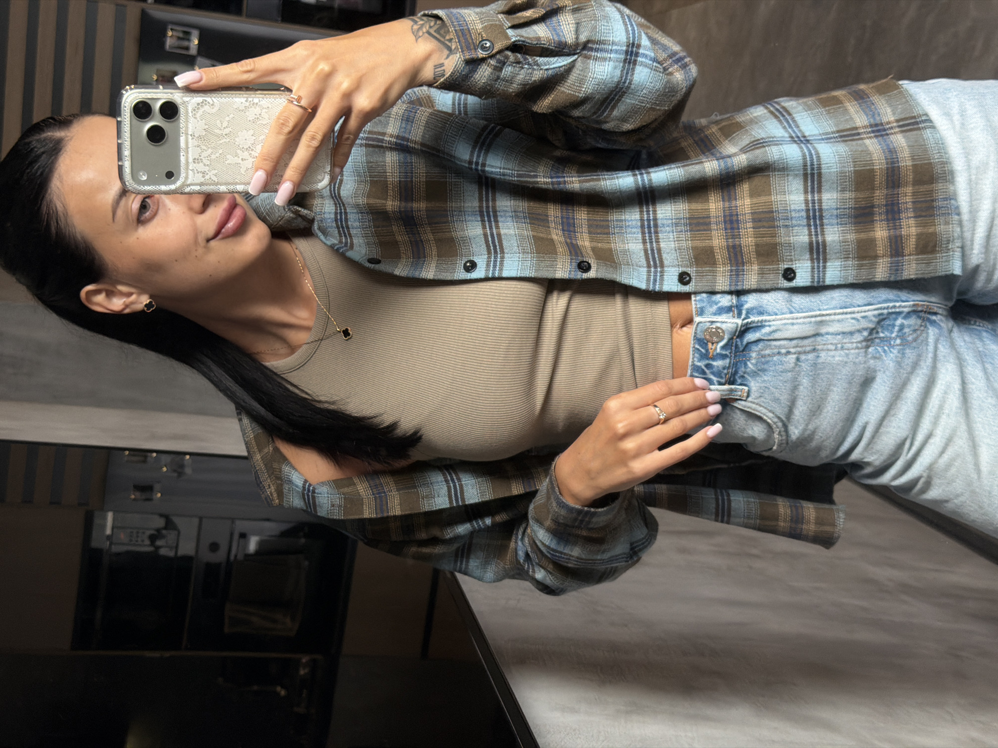A young woman stands in a dimly lit room, holding her phone up to take a selfie. She is wearing a tan ribbed tank top under an open, blue and brown plaid flannel shirt. Her light-wash denim jeans are visible from the waist down, with a silver button and a black button at the fly. She has long, dark hair pulled back and is wearing a small gold necklace with a black clover pendant and a simple gold ring on her right hand. Her left hand is resting on the waistband of her jeans, with her phone in her right hand. The background is out of focus but appears to be a kitchen area with dark cabinets and appliances.