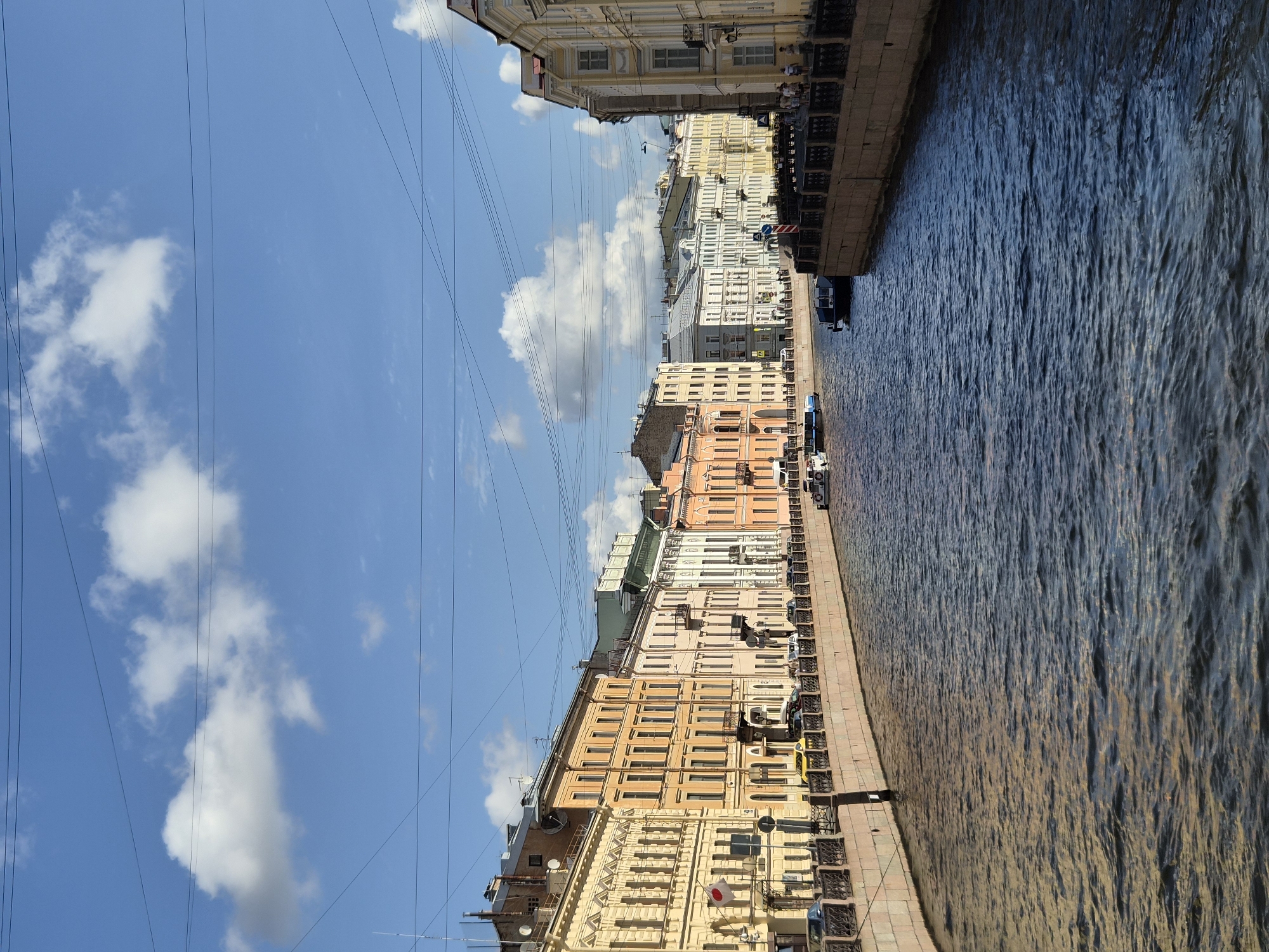The image depicts a wide canal with dark, rippling water reflecting the sky and buildings. On both sides of the canal are rows of multi-story buildings with classical architecture, painted in pastel colors like yellow, orange, pink, and white. The buildings feature numerous windows, balconies, and decorative details. A stone embankment with an ornate metal railing lines the canal, and a street with parked cars and a few pedestrians runs behind it. Several boats are visible on the water, including a small tour boat. The sky above is bright blue with scattered white, fluffy clouds, and overhead power lines crisscross the scene. The overall impression is of a vibrant city canal on a sunny day.