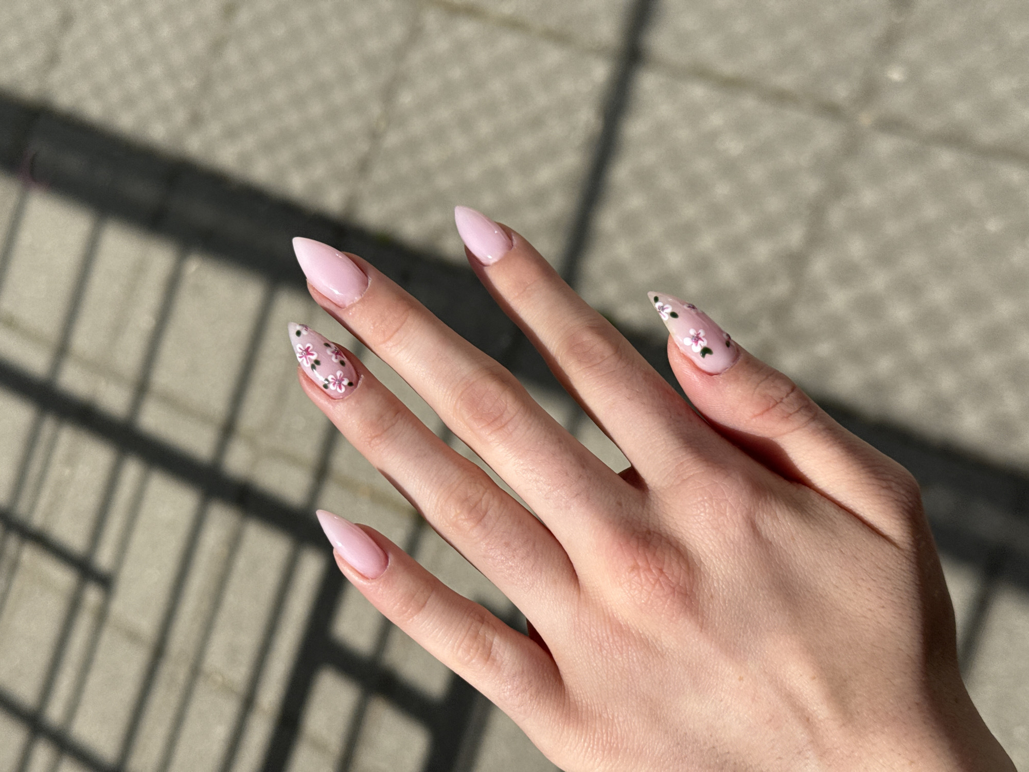 A close-up shot features a light-skinned hand, likely female, showcasing its meticulously manicured nails against a sunlit, outdoor background. The nails are long and shaped in a stiletto or almond style, painted in a delicate, light pink polish. Two nails, the ring finger and pinky finger, are adorned with intricate floral designs. These patterns consist of small white and pink flowers accented with green leaves and dark dots, adding an elegant detail to the otherwise monochromatic pink set. The thumb, index, and middle fingers are painted solely in the soft pink hue.
The hand is positioned against a textured, light gray ground surface, possibly concrete or pavement. Strong, defined shadows of an overhead structure, resembling a grid or parallel lines, are cast across the background, indicating bright daylight conditions, likely late morning or afternoon. The clear shadows suggest sunny weather. There is no visible text in the image. The scene is set in Plovdiv, Bulgaria.