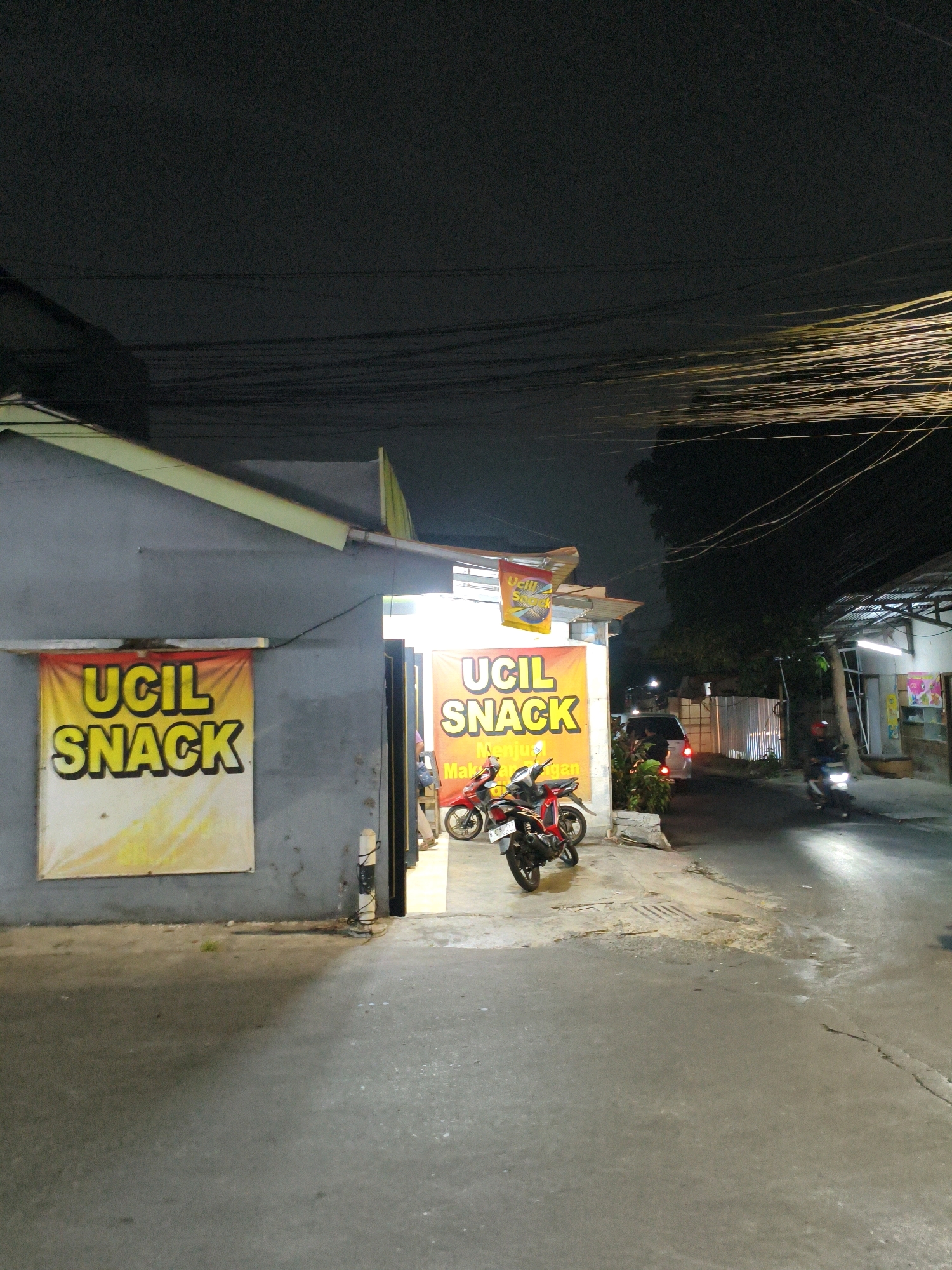 This image depicts a street scene at night in Bekasi, Indonesia. The foreground features a paved road with a slight sheen, suggesting it may be wet. To the left, a gray building has a large yellow banner with the text "UCIL SNACK" in bold black letters. Another similar banner is visible to the right, also advertising "UCIL SNACK." Several motorcycles are parked in front of the building. One red motorcycle is prominently displayed, and another is partially visible behind it. A person can be seen standing near the entrance of the establishment. Further down the street, a white van is parked, and its brake lights are illuminated. A person is operating a motorcycle on the right side of the street, its headlight casting a bright beam. The background is dark, with the silhouettes of trees and buildings against the night sky, and numerous overhead wires crisscross the upper portion of the image. The overall impression is of a commercial area at night, with a focus on a snack vendor.