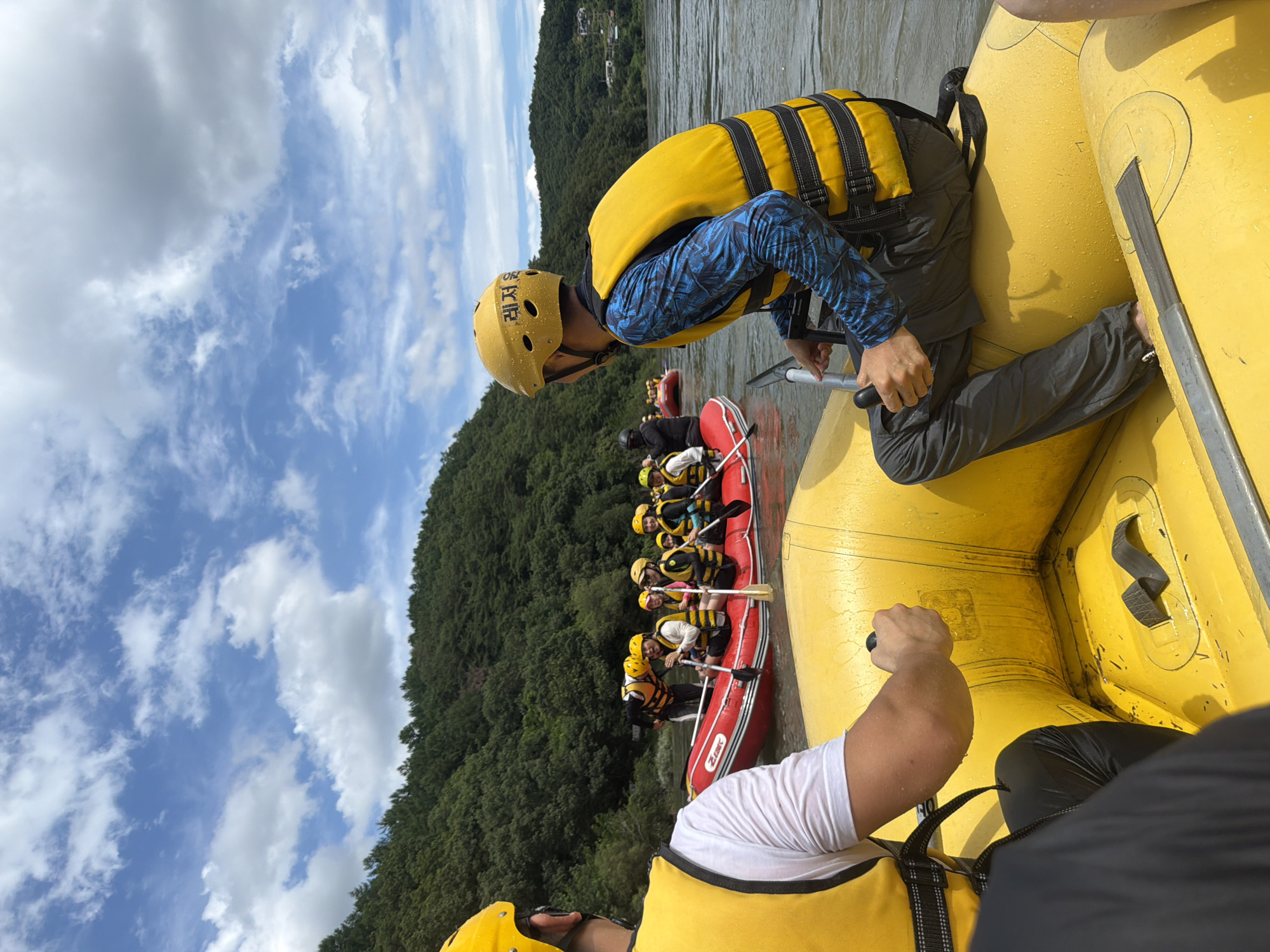 The image depicts a group of people participating in a white-water rafting activity on the Geumgang River in Seo-myeon, South Korea. The perspective is from within a yellow inflatable raft, looking towards another red inflatable raft carrying several individuals. Both rafts are equipped with paddles, and the participants are wearing yellow safety helmets and bright yellow life vests. The people in the red raft are actively paddling, suggesting movement through the water. The environment is characterized by a wide, calm river with a backdrop of lush green, tree-covered hills and a partly cloudy sky. The overall scene conveys a sense of outdoor recreation and adventure.
