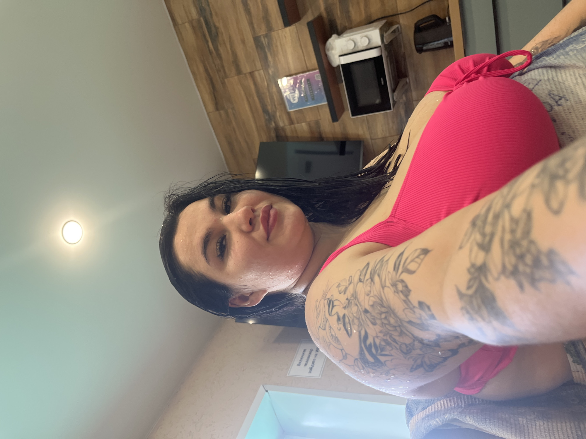 The image captures a selfie of a young woman from a low-angle perspective, suggesting she is lying down and looking up at the camera. She has long, dark, wet hair and is wearing a bright pink ribbed bikini top. Her left arm and shoulder are adorned with extensive black-and-grey tattoos. Her skin has a glistening appearance, suggesting she may have recently been in water. She displays a relaxed expression with slightly parted lips.
The background features a light-colored ceiling with a recessed circular light fixture on the upper left. To her right, a wall with horizontal light wood paneling is visible. On a surface below the wooden wall, a small white microwave or compact oven, and a dark flat-screen object (likely a TV) can be seen, along with some packaged items. A patterned towel or blanket is partially visible under her right arm.
A small white sign with black text in Cyrillic is affixed to the wooden wall. The visible text reads "Включите телевизор" (Turn on the TV), and partially "забудьте на W" (forget about W or log on to W... likely referring to Wi-Fi).
The indoor setting, wet appearance, and presence of appliances and a TV instruction sign suggest a hotel room, spa area, or changing facility. The time of day is not determinable, as the lighting is artificial.