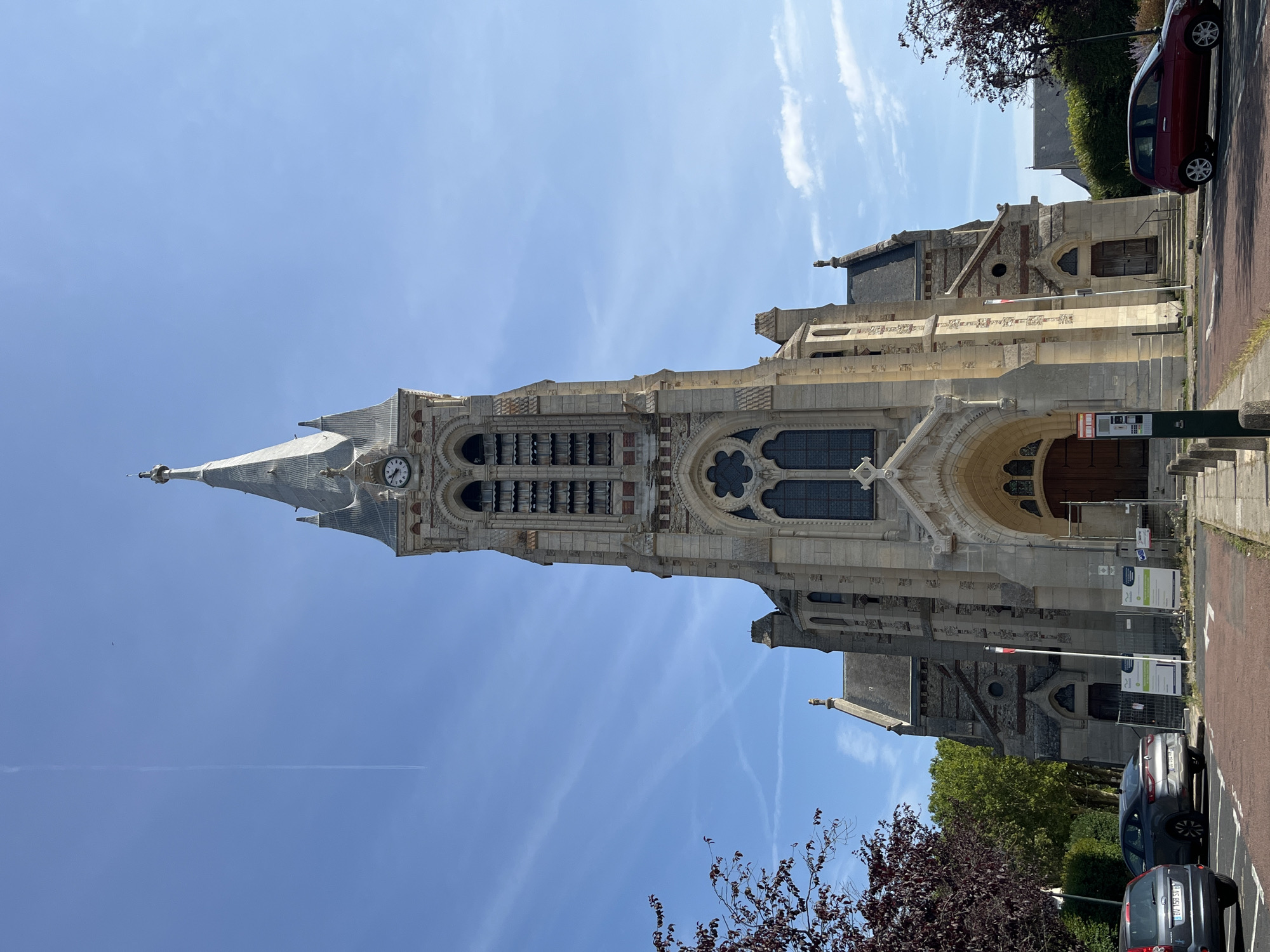 The image shows the facade of the Saint-Lubin church in Rambouillet, France, on a clear day with a blue sky and some wispy clouds. The church is constructed of light-colored stone and features a tall, ornate bell tower with a spire and a clock face. The main entrance is a large arched doorway with wooden doors. To the left and right of the main entrance are smaller arched doorways. There are trees with dark red leaves on the left side of the image and trees with green leaves on the right. Several cars are parked in a paved area in front of the church, and some temporary construction barriers and signs are visible. A red car is driving away from the church on the right side of the image.