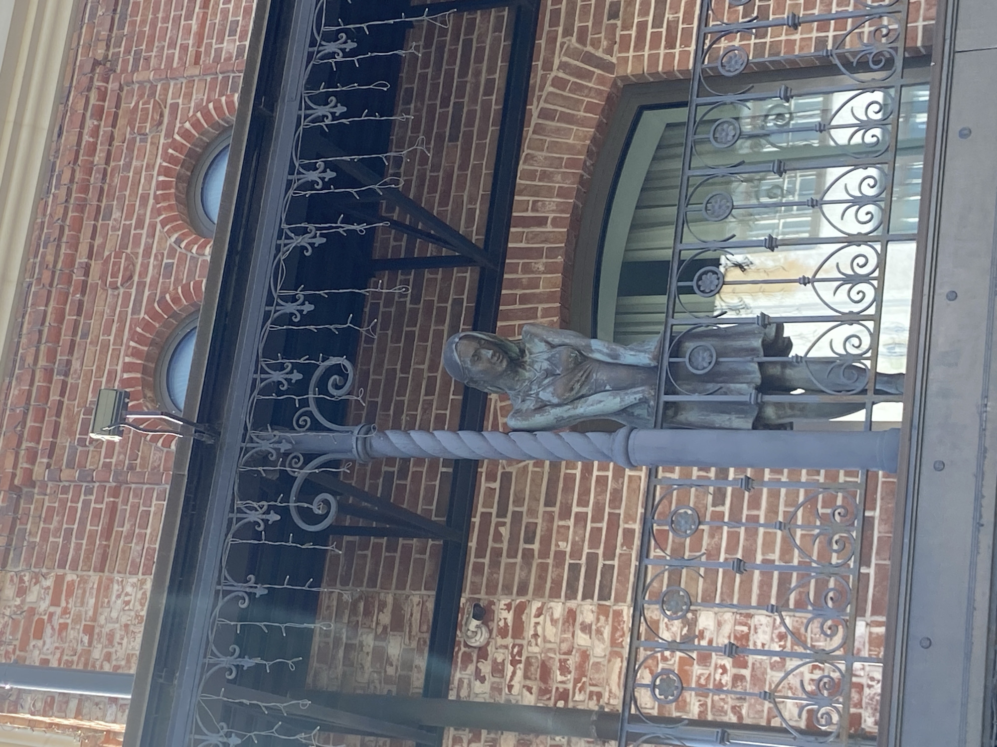 A bronze statue of a woman, possibly a decorative element, is mounted on a decorative wrought-iron structure, likely a gate or balcony railing. The statue appears to be of a woman in repose or contemplation, with draped clothing. The wrought iron is painted a dark grey and features ornate curls and decorative elements. The background consists of a red brick building with arched windows. The location appears to be in Kazan, Russia, as evidenced by the architectural style of the building and the wrought-iron details. The angle of the shot suggests it was taken from below, looking upward at the statue and wrought iron.