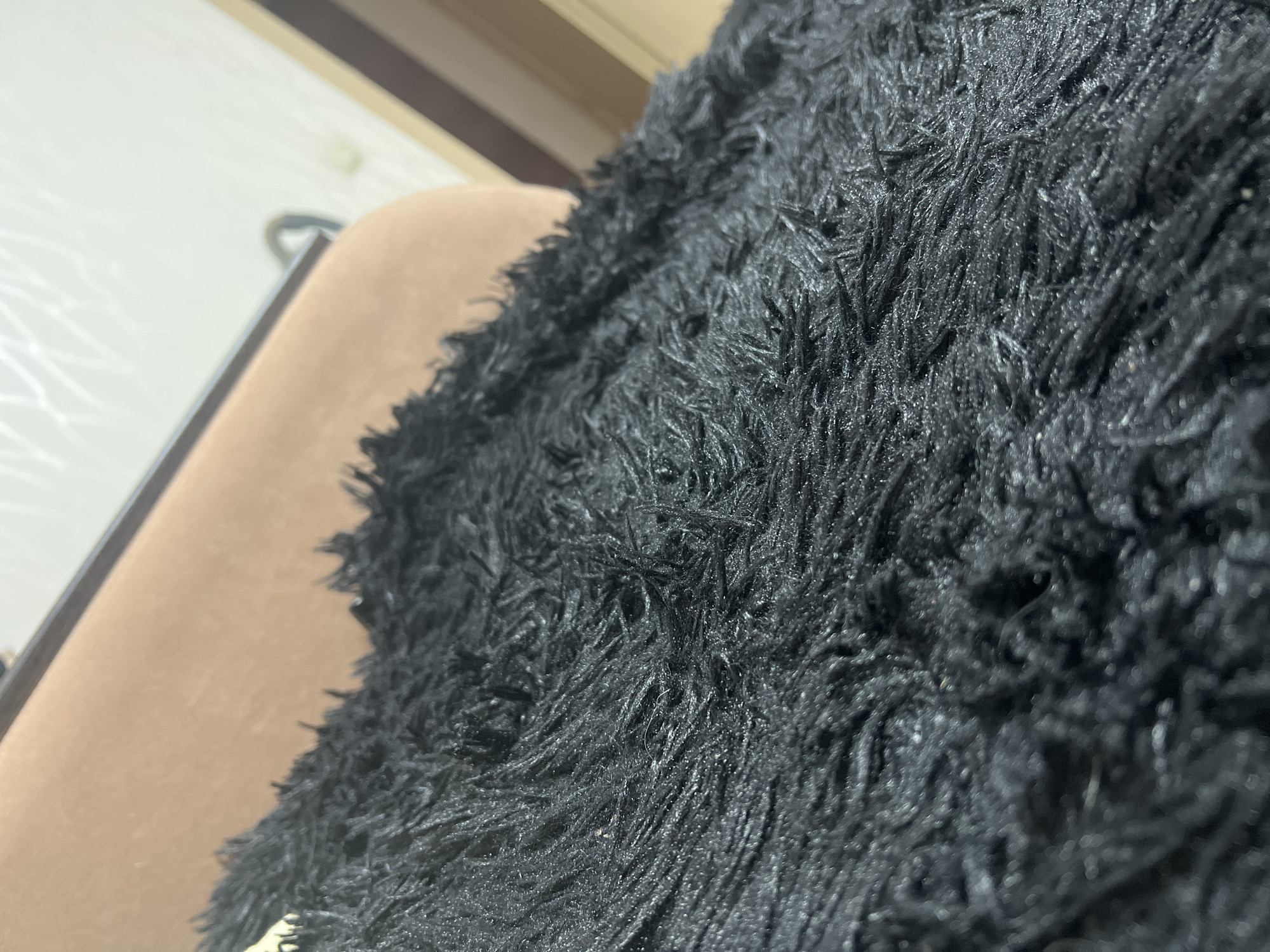 The image shows a close-up of a black, shaggy material resting on a tan, smooth surface. A blurred white surface is visible in the background, along with a partially visible brown vertical stripe. The location of the image is unknown, and there is no information to suggest it was taken in Perm, Russia.