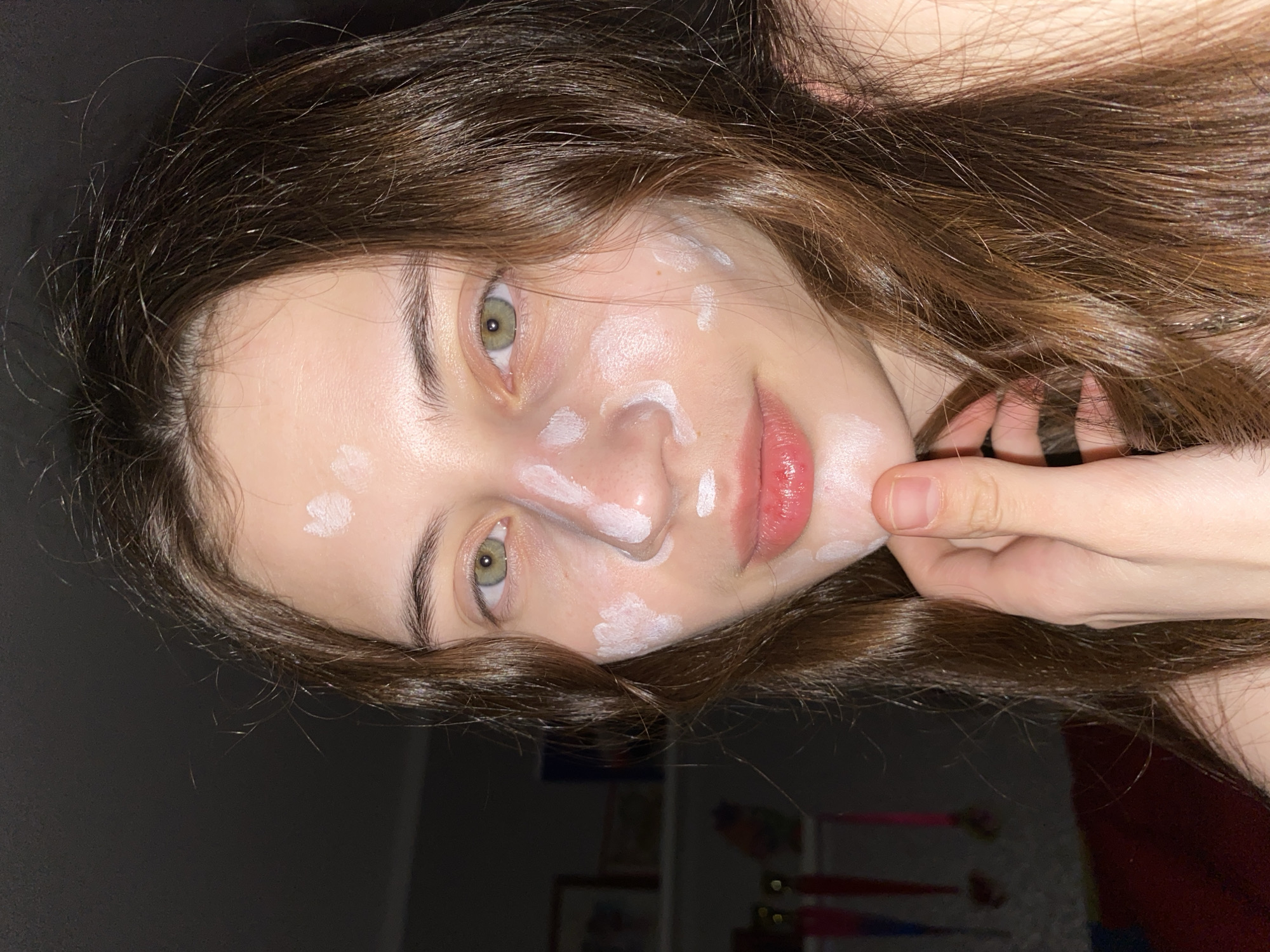 A young woman with long, wavy brown hair and green eyes poses for a close-up selfie. She has small, white dollops of cream applied to her face, mostly on her nose, cheeks, and around her mouth. Her skin appears fair, and her lips are lightly glossy. A hand is visible near her chin. The background is dark, suggesting a dimly lit room. Unidentifiable objects are blurred in the far background. There's no location data to specify the city of Votkinsk, Russia.