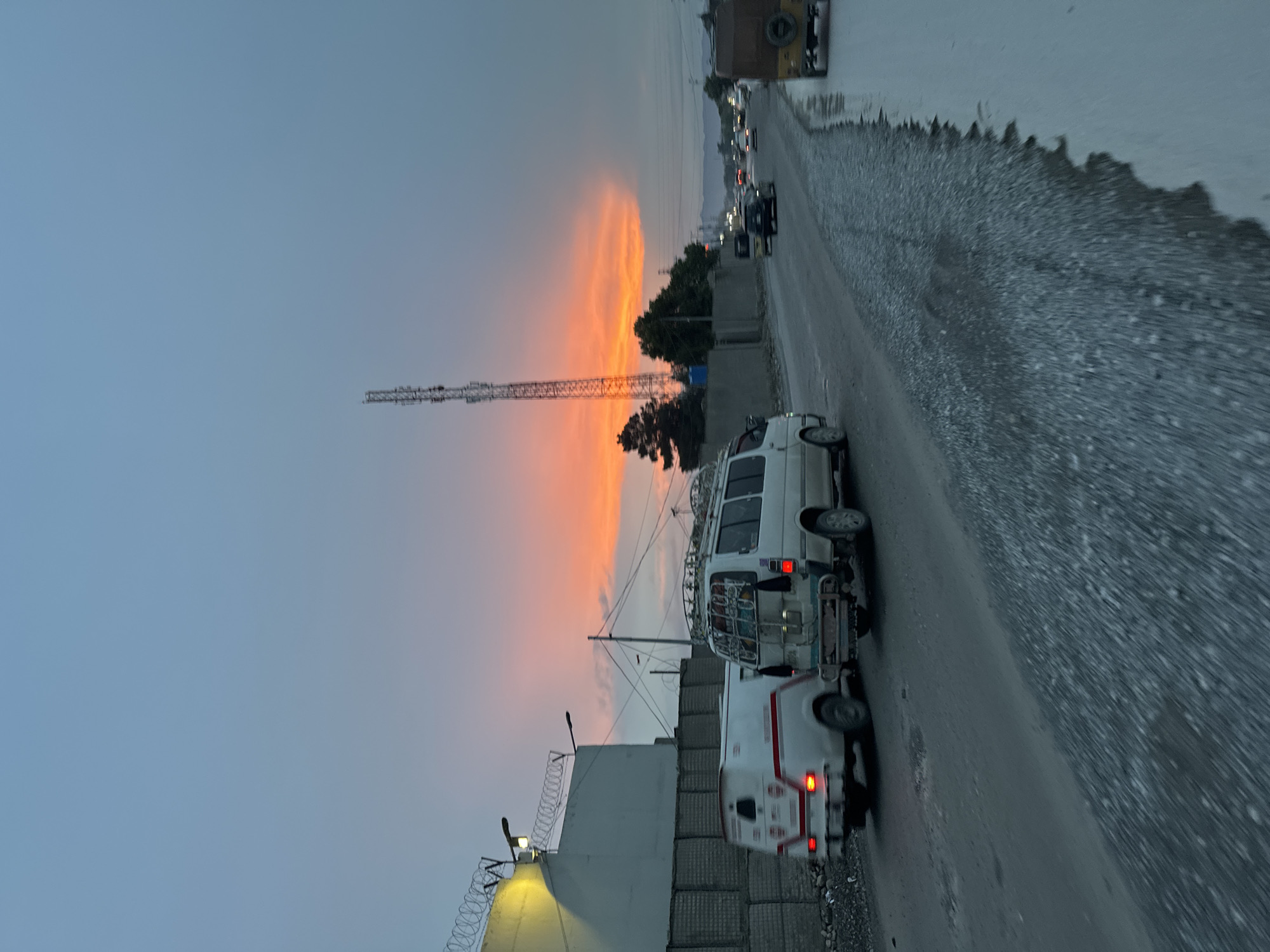 The image depicts an urban scene in Quetta City Tehsil, Pakistan. A gray asphalt road runs through the foreground and background, with loose gravel and debris along the right side. Two vans, a white minibus with a rack on top and a smaller white van with red accents, are parked on the road. In the background, a line of cars and a truck are visible on the road, receding into the distance. A communications tower stands tall in the middle ground, piercing a dramatic sunset sky with orange and pink hues. To the left, a concrete wall topped with barbed wire is partially visible.