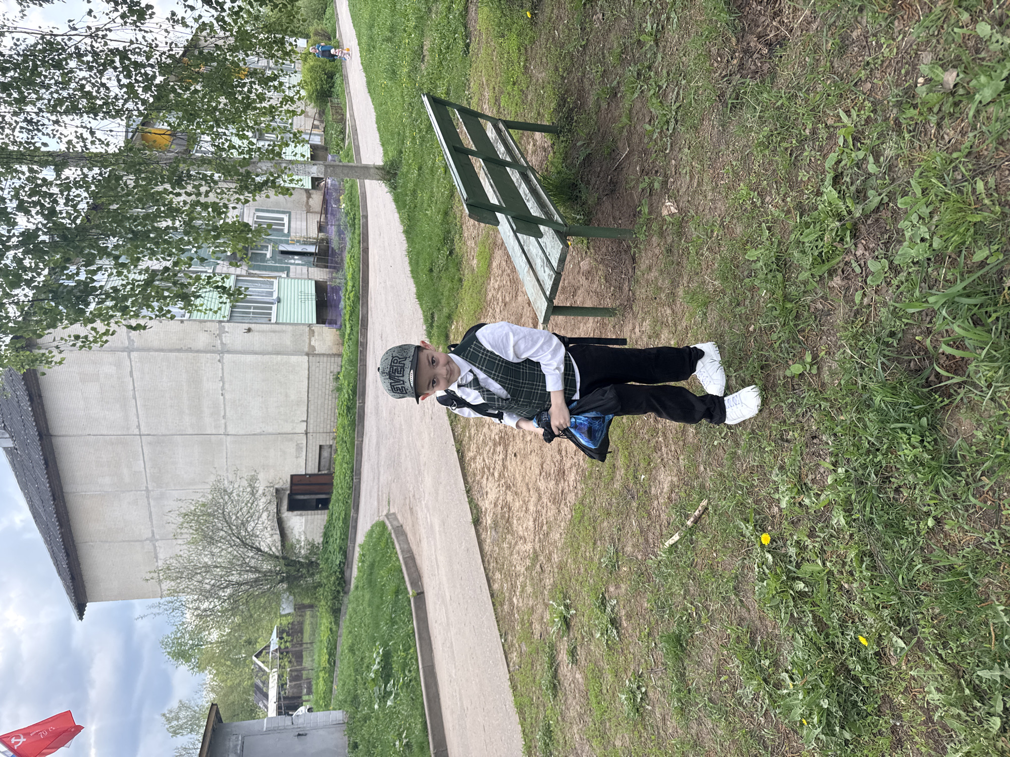 The image depicts a boy standing in a park-like setting in Раздолье, Russia. He is wearing a cap, a white shirt, a plaid vest, black pants, and white sneakers. He is holding a blue bag. Behind him is a green bench and a paved pathway. To the left is a multi-story building. The area consists of grass and sparse vegetation, with a few dandelions. In the background, other buildings and trees can be seen. The sky is overcast.