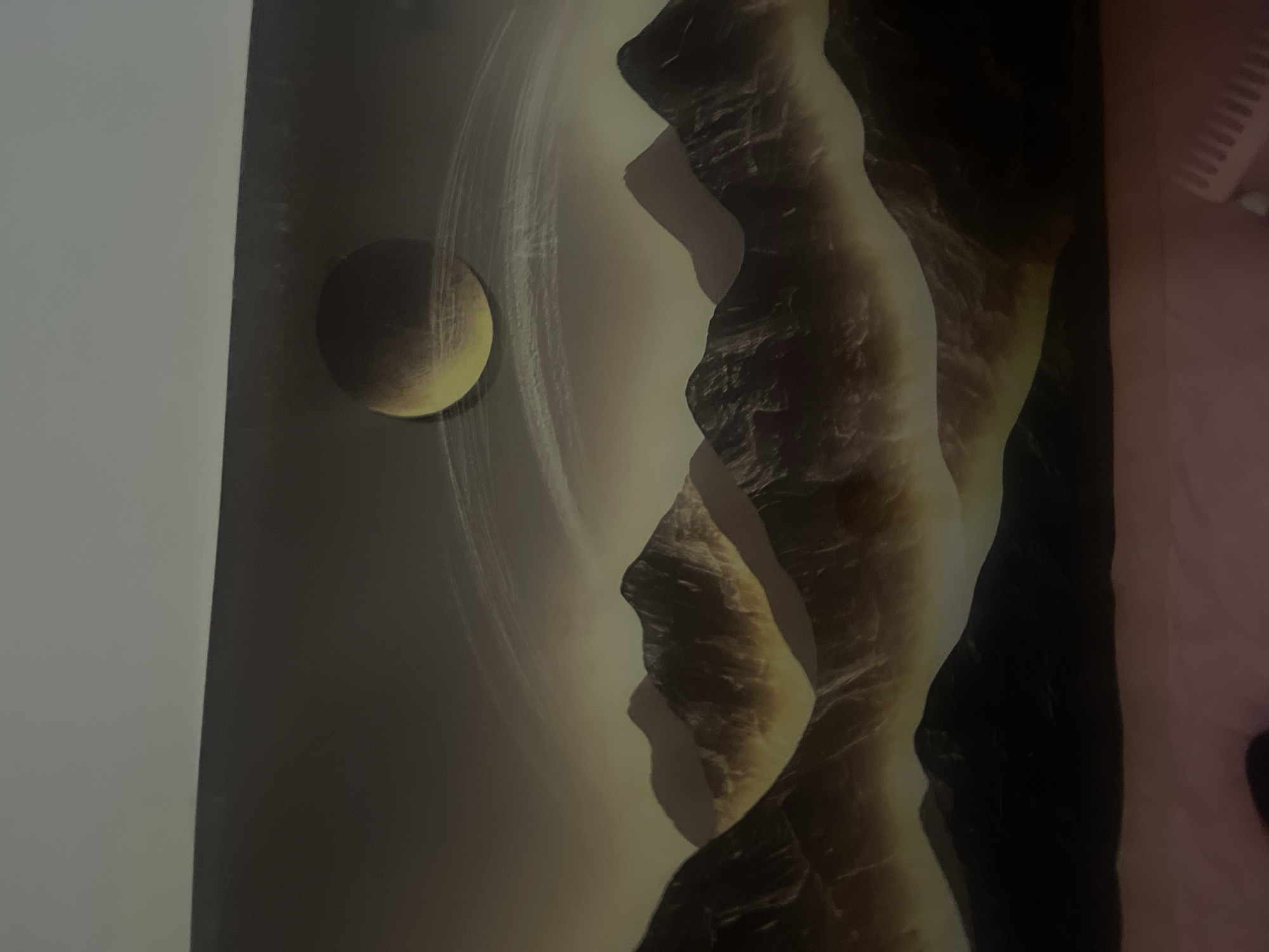 The media file is an image depicting an artistic representation of a planet with rings and a moon. The planet is a sphere with a yellowish-brown hue, surrounded by luminous, ethereal rings that appear to be made of light or dust. To the left of the planet, a smaller celestial body, possibly a moon, is visible. The scene is dominated by a dramatic, rocky landscape that rises vertically from the bottom of the frame, casting deep shadows. The rocks are textured and appear to be illuminated from the side, creating a sense of depth and dimension. The overall color palette is muted, with tones of yellow, brown, black, and a soft, hazy background. The lighting suggests a dramatic, possibly twilight or alien, setting, with strong contrasts between light and shadow. There is no visible text within the image.