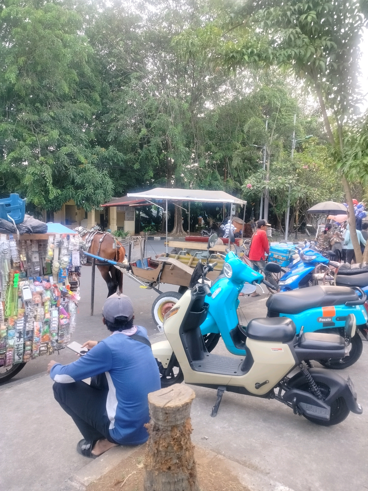 In East Jakarta, Indonesia, a brown horse pulls a wooden cart filled with various items, likely for sale. The cart is positioned next to a street vendor's display of goods, which includes many small, packaged items hanging from racks. A man wearing a blue long-sleeved shirt and black pants is crouched on the ground near the cart, looking at a mobile phone. Several blue electric scooters are parked behind the cart, one of which has the word "Fazzio" visible on its side. In the background, there are trees and buildings, suggesting an outdoor market or street scene. The overall environment appears to be a public area with both traditional and modern forms of transportation present.