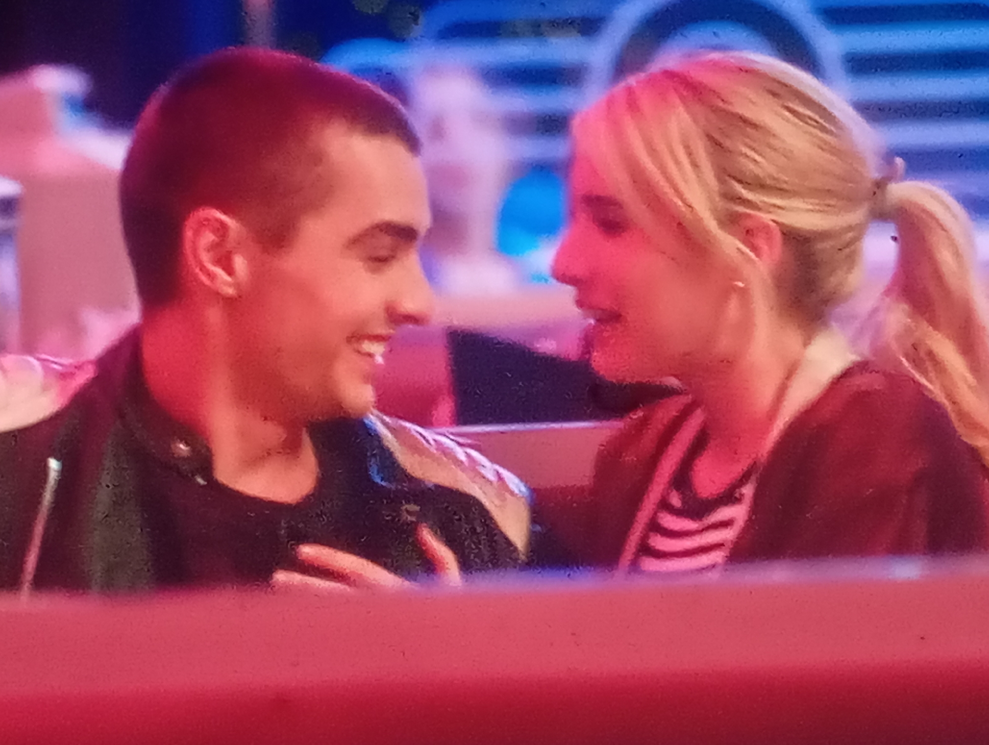 The image is a close-up, slightly blurry shot of a man and a woman in what appears to be a dimly lit indoor setting. The lighting is predominantly red and blue. The man, on the left, has short dark hair and is looking to his right, smiling. He is wearing a dark jacket with some lighter-colored panels on the shoulders. The woman, on the right, has blonde hair pulled back in a ponytail and is looking at the man. She is wearing a dark top with horizontal stripes. The background is out of focus, showing blue and indistinct shapes. There is no indication of Sofiivska Borshchahivka, Ukraine in the image.
