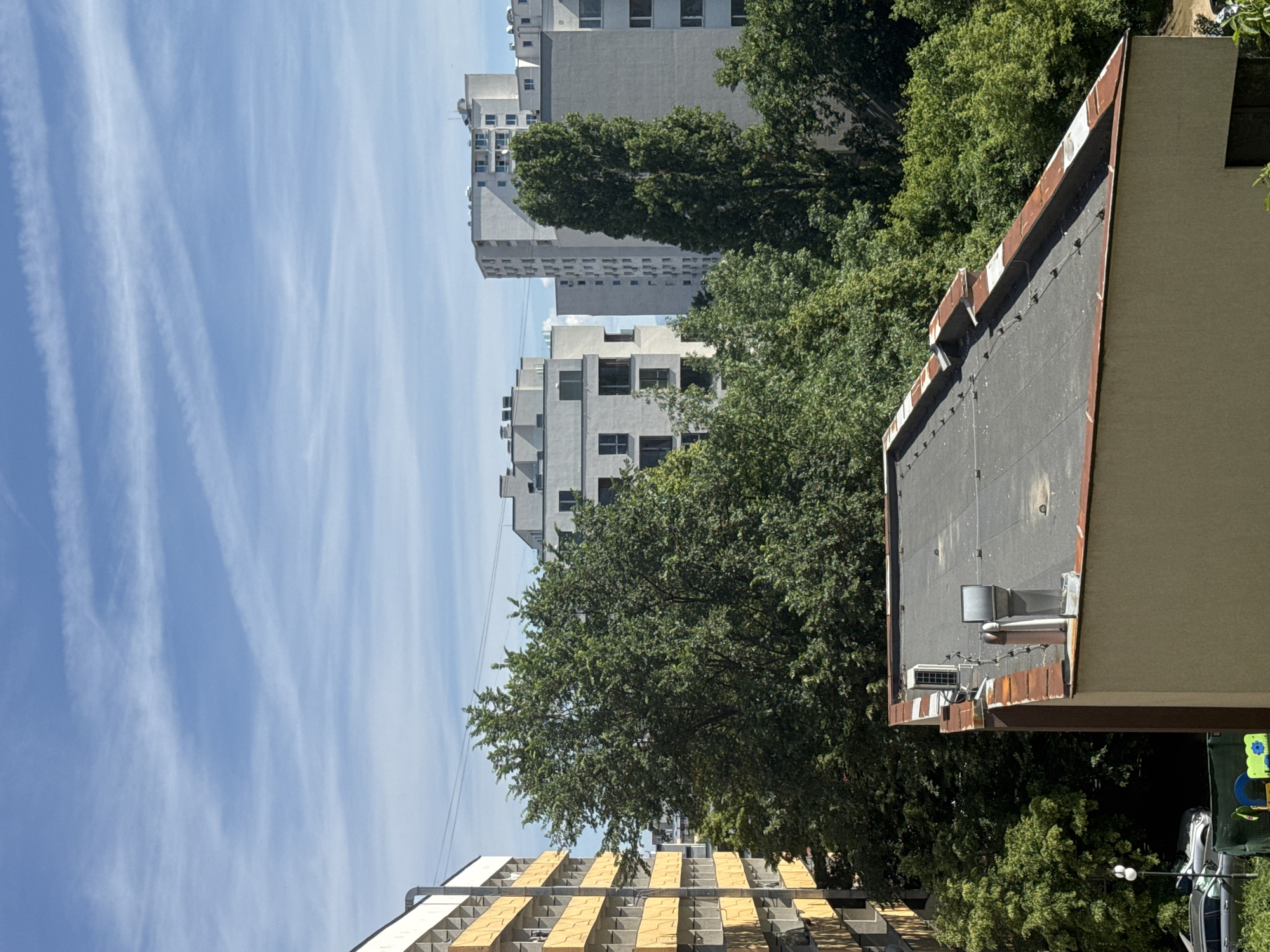 An aerial view captures the urban landscape of Sofia, Bulgaria, on a bright, sunny day with a partly cloudy sky. In the foreground, the flat, dark-grey roof of a building with a reddish-brown trim is prominent. Two vent pipes and an air conditioning unit are visible on the roof, along with several small metal fixtures.
To the left, a building with a yellow facade and repeating grey balcony structures is partially visible. Behind these structures, a dense canopy of green trees dominates the midground, with a few taller, slender trees interspersed among the broader foliage.
In the background, several modern, multi-story apartment buildings with white facades rise above the trees. These buildings feature numerous windows and balconies. The sky above is a vibrant blue, streaked with thin, white cirrus clouds.
At the lower left of the image, two cars are parked on a street, and a small children's playground structure can be seen. Power lines are strung across the scene, connecting some of the buildings. The overall impression is one of a residential urban area bathed in daylight.