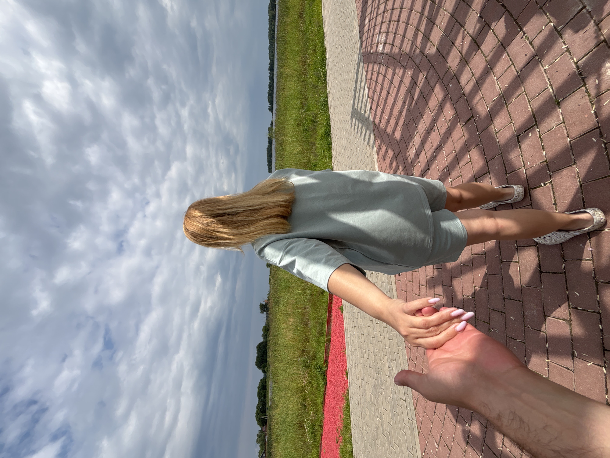 A first-person perspective shot shows a person's hand holding another person's hand as they walk along a brick-paved path. The person whose hand is being held is facing away from the camera and is wearing a light blue t-shirt and shorts set with patterned slip-on shoes. Their blonde hair is visible. The path is curved and casts a shadow pattern on the ground. Beyond the path, there is a strip of red gravel followed by a field of tall green grass and reeds. In the distance, there is a body of water, trees, and a cloudy sky. The location is specified as Breytovo, Russia.
