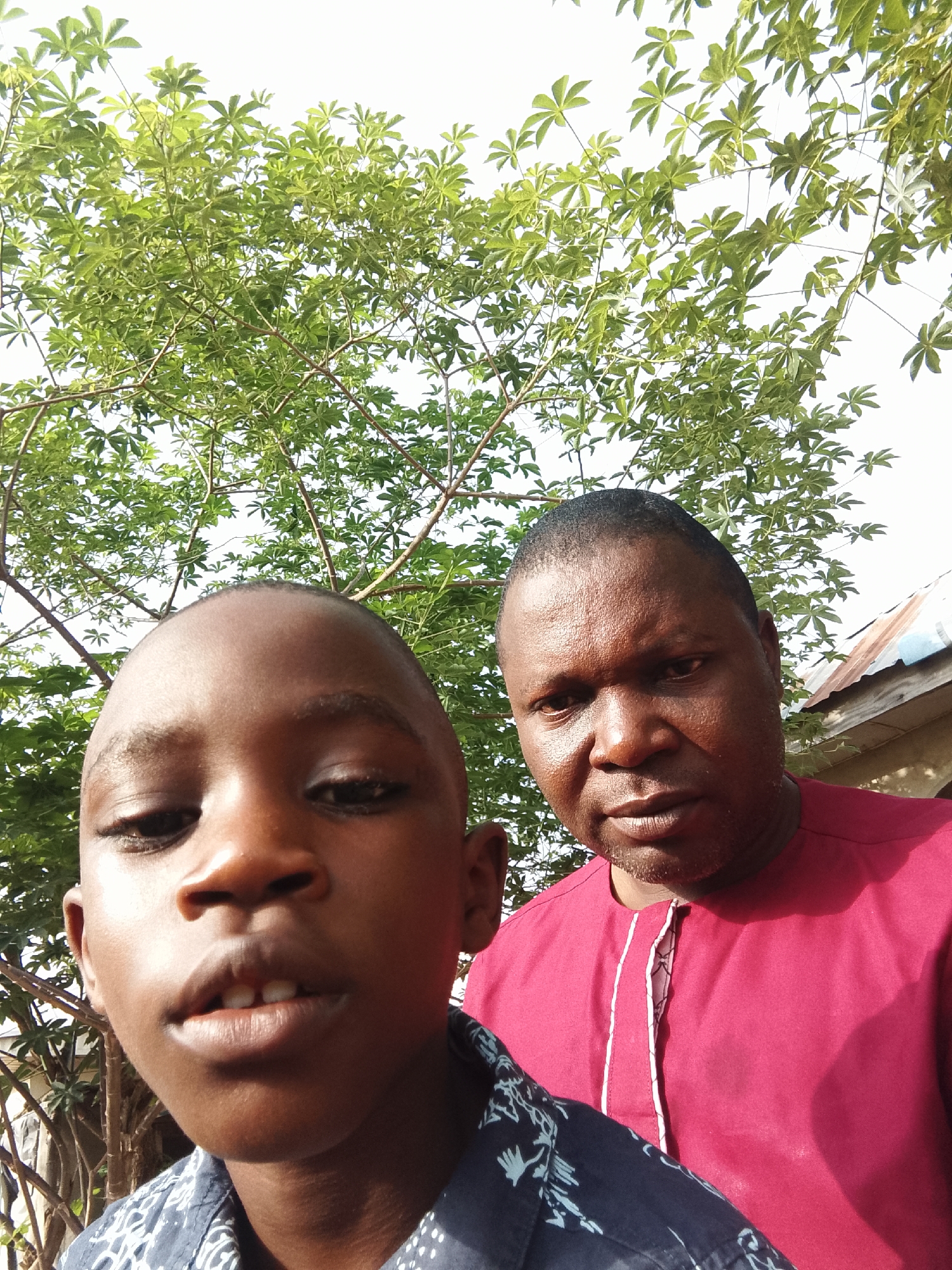 A medium shot shows two people, a young boy and an older man, posing for a selfie. The boy is in the foreground, while the man is positioned slightly behind him to the right. Lush green foliage fills the background, with tree branches and leaves visible above them. A glimpse of a building structure can be seen partially on the right side, with a portion of its rusted roof apparent. The city of Unknown, Unknown is not identifiable in this image.