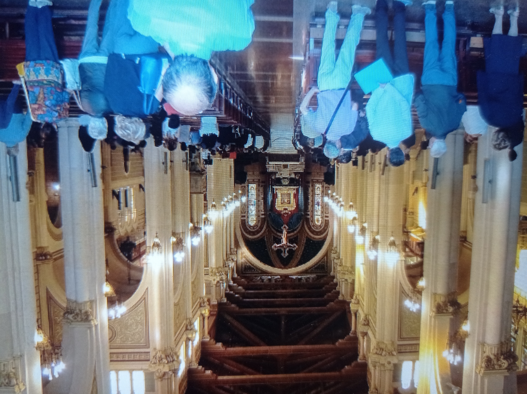 The image shows the interior of a large, ornate cathedral, likely in the city of Mukachevo, Ukraine, given the context. The architecture features high, vaulted ceilings with wooden beams, tall, fluted columns with decorative capitals, and arched alcoves with lighting. At the far end of the nave, a prominent crucifix hangs above an altar area, framed by arched windows.
Several people are visible throughout the cathedral, some standing and facing towards the altar, while others are seated in the pews. The individuals are dressed in casual attire, suggesting a visit or a service is in progress. The lighting appears to be a mix of natural light from the high windows and artificial lights mounted on the columns and within the architecture, creating a warm, illuminated atmosphere. The floor is reflective, suggesting it is polished stone or tile. The overall impression is one of a grand, sacred space filled with worshippers.