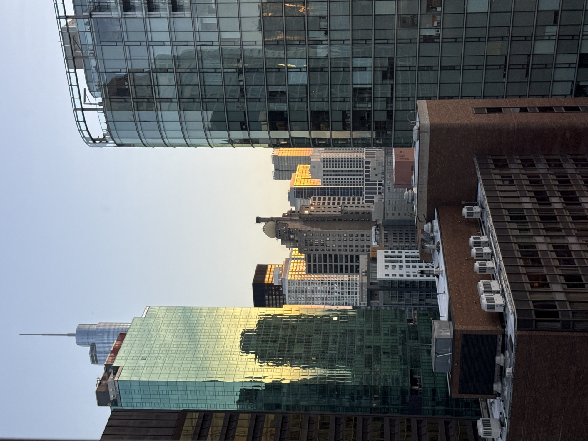 This is a photograph of a cityscape, likely taken from an elevated position, showcasing a collection of modern high-rise buildings under a clear, pale blue sky. The buildings are constructed primarily of glass and steel, with varying architectural styles and heights.
On the left, a tall, slender building with a pointed spire pierces the sky, its upper section clad in a reflective, light-colored material. Adjacent to it, a striking green glass skyscraper dominates the middle ground, its facade reflecting the surrounding structures and the warm glow of what appears to be the setting or rising sun. The reflection on this building shows distorted silhouettes of other buildings.
To the right, a curved, modern glass skyscraper with a rounded corner occupies a significant portion of the frame. Its exterior is composed of numerous glass panes, and the interior spaces are visible, suggesting it is occupied. A glass railing is visible along the top of this building, where a few figures can be discerned.
In the foreground and mid-ground, several other buildings of varying heights and architectural styles are visible. Some are clad in brick, while others have darker glass facades. The rooftops of some of these buildings are visible, revealing HVAC units and other rooftop equipment. The overall scene is one of urban density and contemporary architecture. The light suggests it is either early morning or late afternoon, casting long shadows and highlighting the reflective surfaces of the buildings.
Without additional information or context, pinpointing the exact location within Chicago, United States of America, is not possible solely from the visual cues in the image. However, the style of architecture and density are consistent with business and residential districts in major American cities.