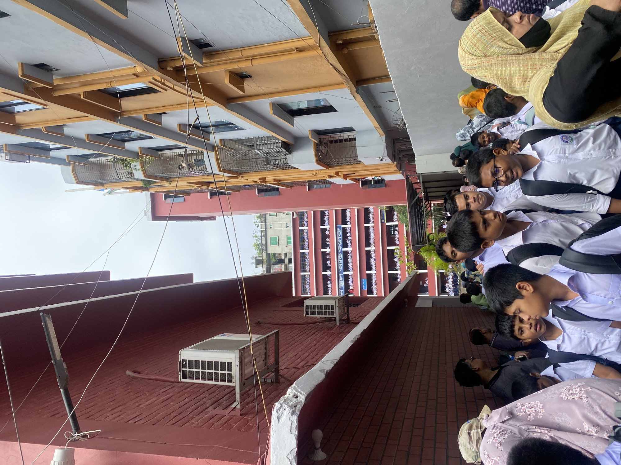 The image shows a narrow alleyway between two multi-story buildings in Dhaka, Bangladesh. The building on the left is a red brick structure with an air conditioning unit mounted on the exterior wall. The building on the right is a modern, multi-story building with orange and grey facades, featuring multiple windows and balconies. A crowd of people, primarily young students in white uniforms and backpacks, are walking down the alley. Some adults are also present, including a woman wearing a headscarf and a mask. The sky is overcast. The scene suggests a school environment or a busy street with pedestrian traffic.