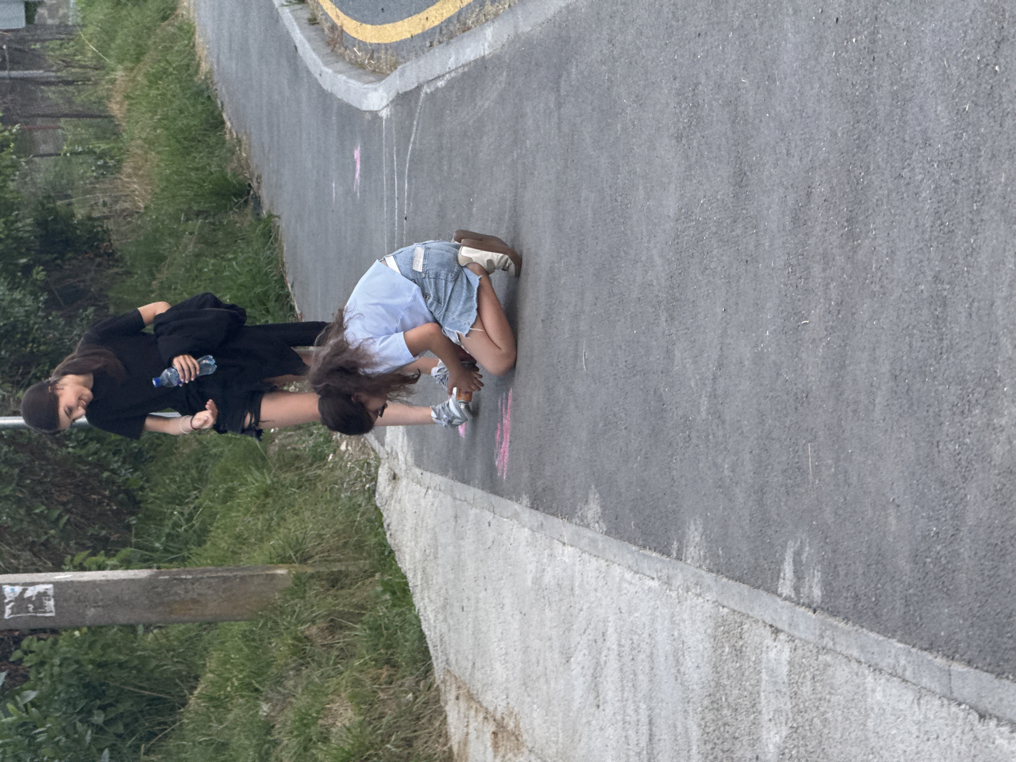 In Sochi, Russia, two young females are observed in the mid-ground of the image. One, positioned in the background, stands with a water bottle and a dark jacket draped over her arm, looking down and smiling. The other, in the foreground, kneels on an asphalt pathway, actively drawing on the ground with chalk. She is wearing a light blue t-shirt, a denim skirt, and sneakers. The environment appears to be an outdoor pathway bordered by grassy slopes on the left and a concrete curb with yellow markings on the right. The pathway itself has chalk markings, some of which are pink. A wooden post with peeling stickers stands to the left of the frame, near the grassy area.