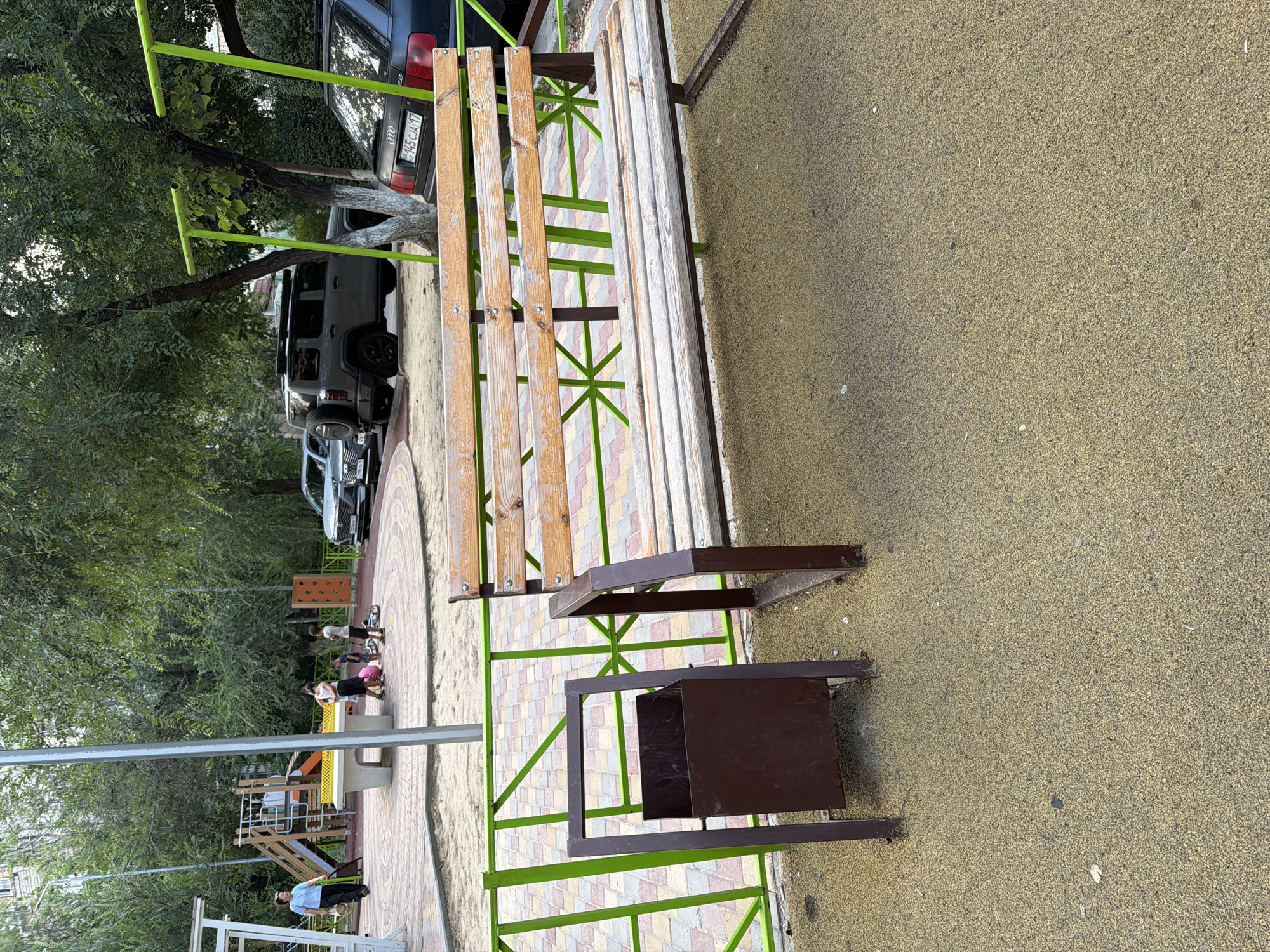The image depicts an outdoor recreational area, likely a playground or park, in Shymkent, Kazakhstan. In the foreground, a wooden bench with green metal supports is prominent. Adjacent to the bench is a dark brown metal trash receptacle. The ground is covered in a textured, yellowish-tan material, possibly rubberized safety surfacing.
In the middle ground, a paved area with a patterned design can be seen, leading towards a playground. Several children and adults are present in this area, some appearing to be engaged in activities on the playground equipment, while others are walking or standing. The playground features slides and other structures.
The background is dominated by lush green trees, suggesting a well-treed environment. Behind the trees, several vehicles are parked, including a dark-colored SUV and another car. One vehicle has a license plate with text that appears to read "145 CJA 17". The lighting suggests it is daytime, with bright but diffused light, indicating possibly overcast or partially cloudy weather. The overall impression is a lively community space utilized by people of various ages.