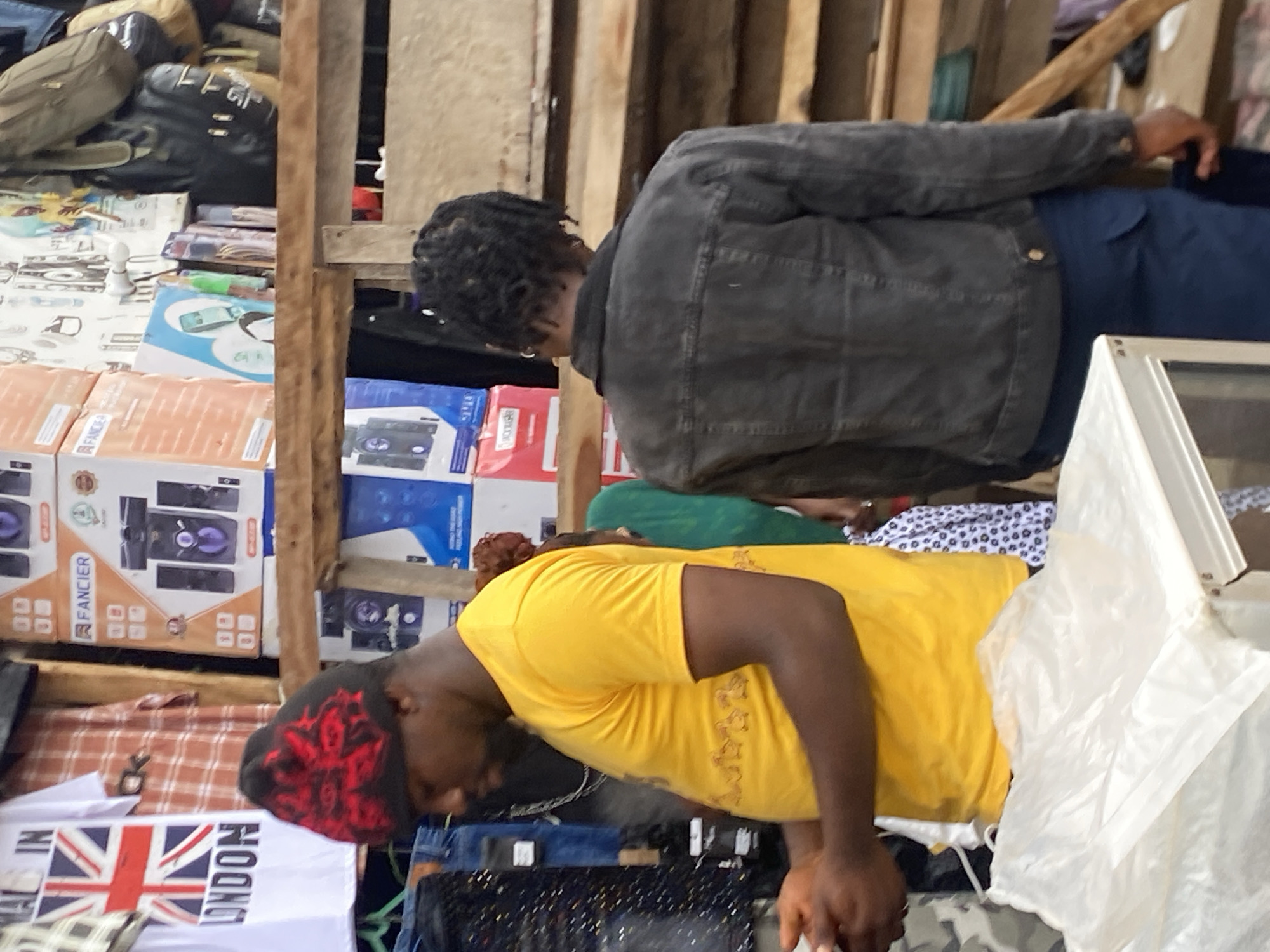 In Bukuru, Nigeria, an outdoor market scene unfolds. A man wearing a yellow t-shirt and a black and red beanie is seen from the side, with his hands clasped in front of him. He stands next to a stall displaying various items. Behind him, another individual in a dark denim jacket and blue pants is also observed from behind. The background reveals shelves stocked with boxes of electronics and bags, suggesting a retail environment. The overall impression is one of bustling commerce in an urban Nigerian setting.