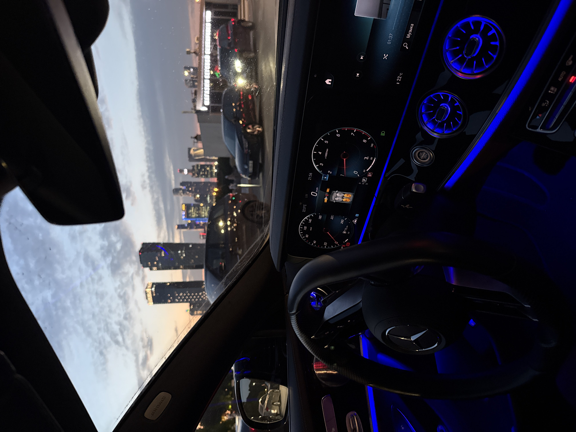 The image is taken from inside a car, looking forward through the windshield and at the dashboard. The car's interior is illuminated with blue ambient lighting. The dashboard displays a digital speedometer and tachometer. Outside the car, a city skyline with illuminated skyscrapers is visible against a twilight sky. Several cars are parked or driving on a road in front of the buildings. One of the buildings has a sign that appears to read "MARKET SECTOR". The overall scene suggests a view of a modern city at dusk from within a luxury vehicle.