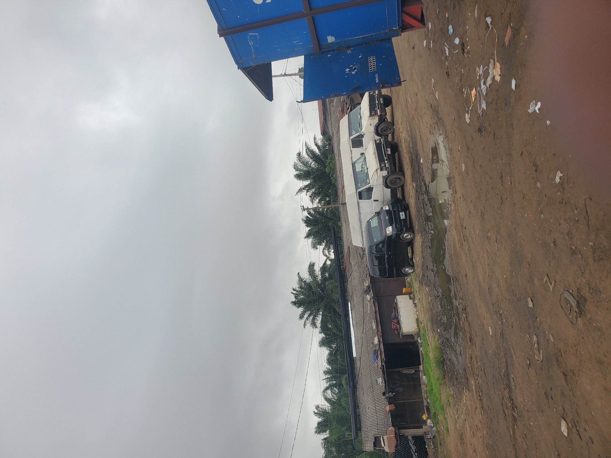 The image depicts a muddy, overcast scene in what appears to be an industrial or commercial area. A row of vehicles, including a black van and several white vans, is parked along a dirt road. A large blue container is situated at the end of the row. Palm trees are visible in the background. Litter is scattered on the ground. The location is consistent with the general characteristics of Fegge, Nigeria, which is known for its commercial activities and diverse infrastructure.