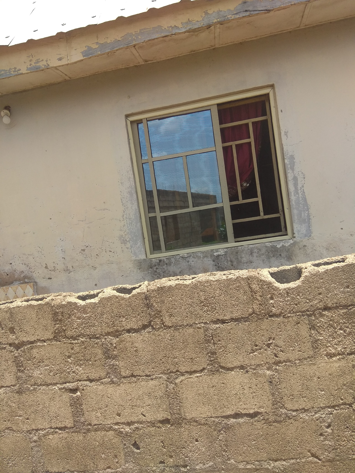 This media file shows a close-up of the exterior of a building and a brick wall in Jalingo, Nigeria. The building wall is a light, faded color with some chipping and discoloration, suggesting age or wear. A single lightbulb fixture is visible on the wall near the top left. A window with a metallic frame is centrally located on the building wall. The window has multiple panes and a metal grate on the inside, behind which a red curtain is visible. Reflections in the window suggest a clear, sunny day with blue skies and some buildings. In the foreground, a textured brick wall made of rough, tan-colored blocks is prominent. The wall is uneven and appears to be constructed with visible mortar. The overall scene depicts an outdoor setting, likely during daylight hours given the lighting and reflections. There are no people visible, and the activity is limited to the static view of the architecture.